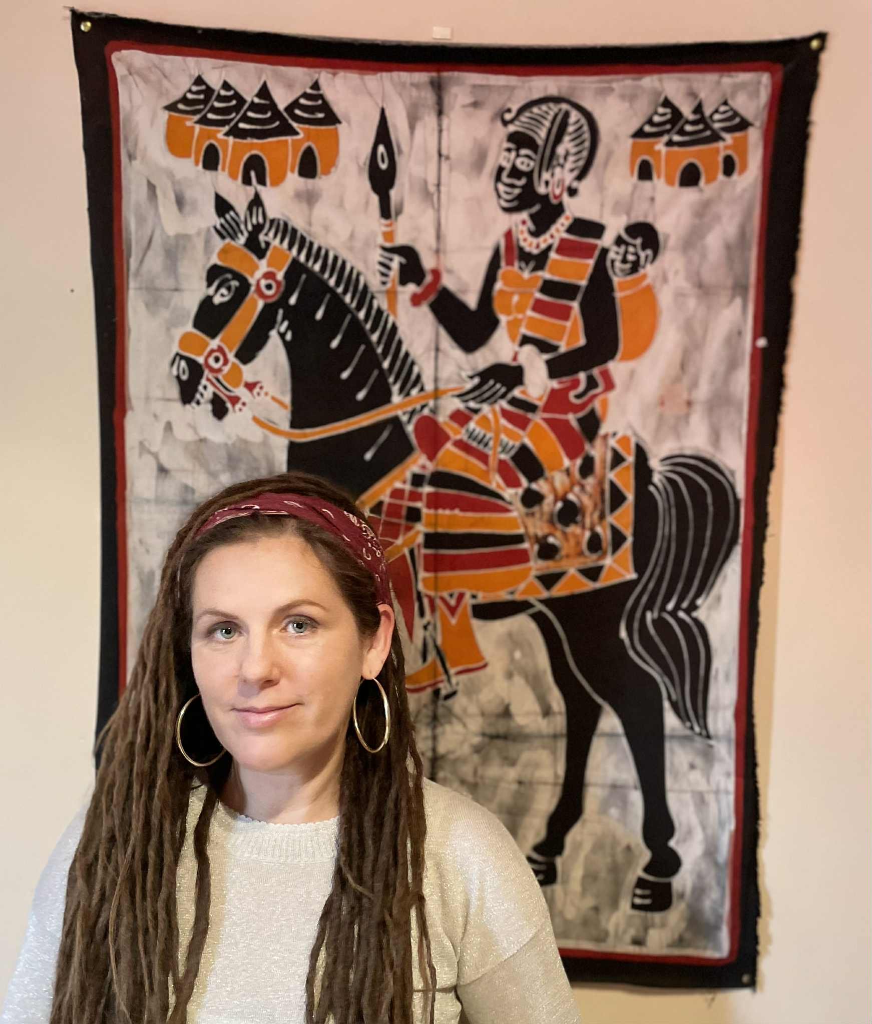 A woman with long dreadlocks and hoop earrings standing in front of a colorful wall hanging depicting a detailed African-style illustration of a woman, a horse, and traditional huts.
