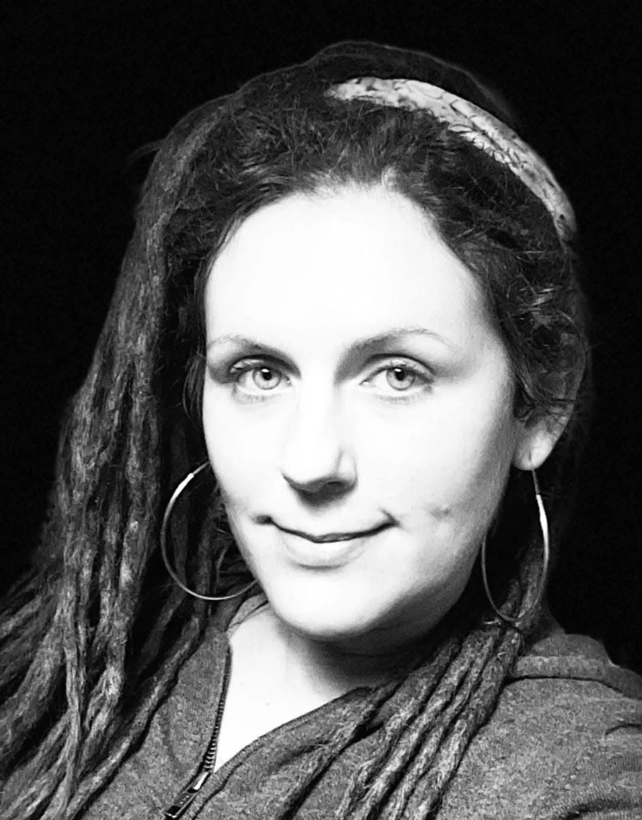 Black and white portrait of a woman with light-colored eyes, long dreadlocks, wearing hoop earrings and a zip-up jacket, smiling slightly.