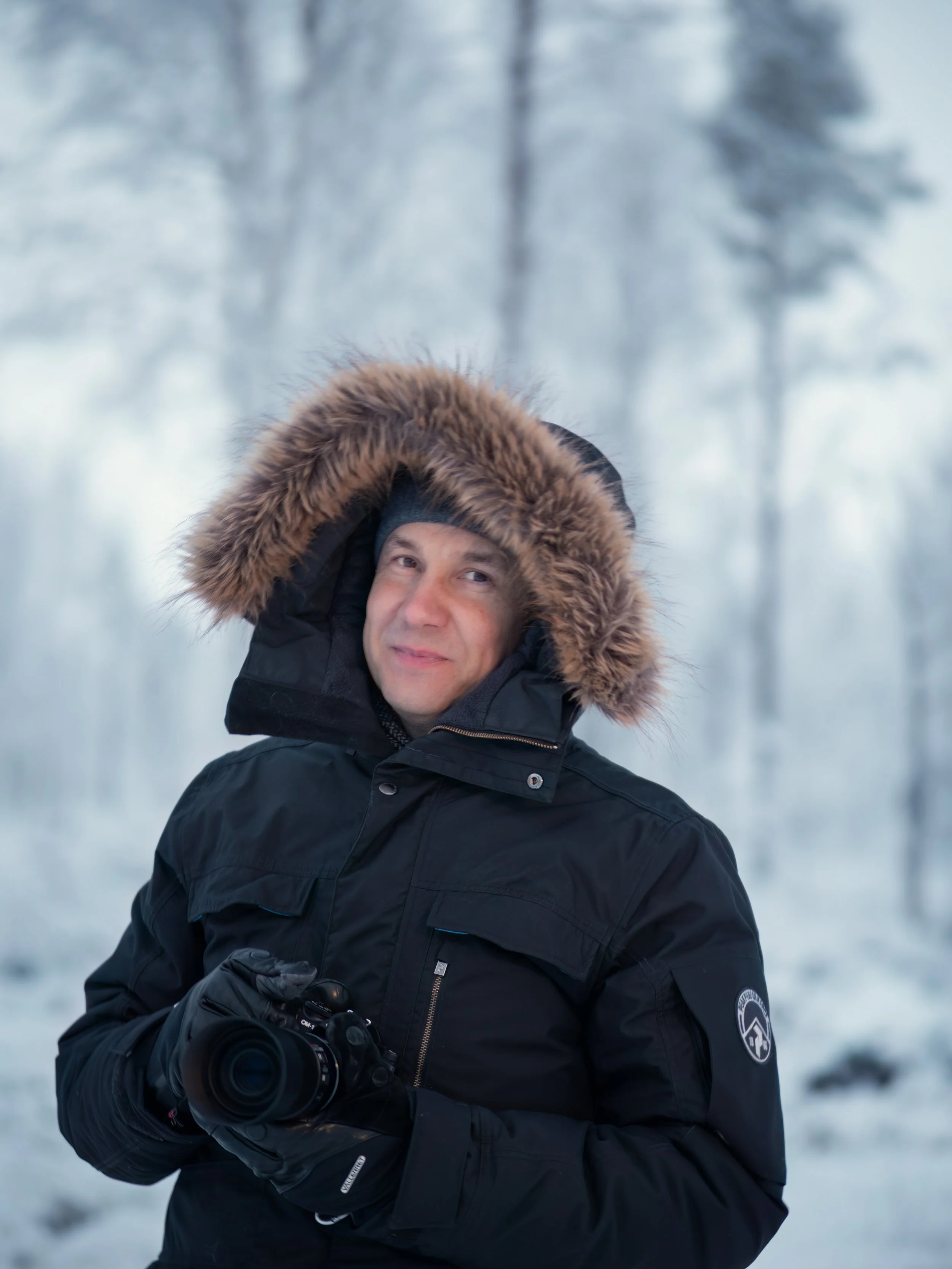 A man in a black winter jacket with a fur-lined hood holding a camera outdoors in a snowy forest.