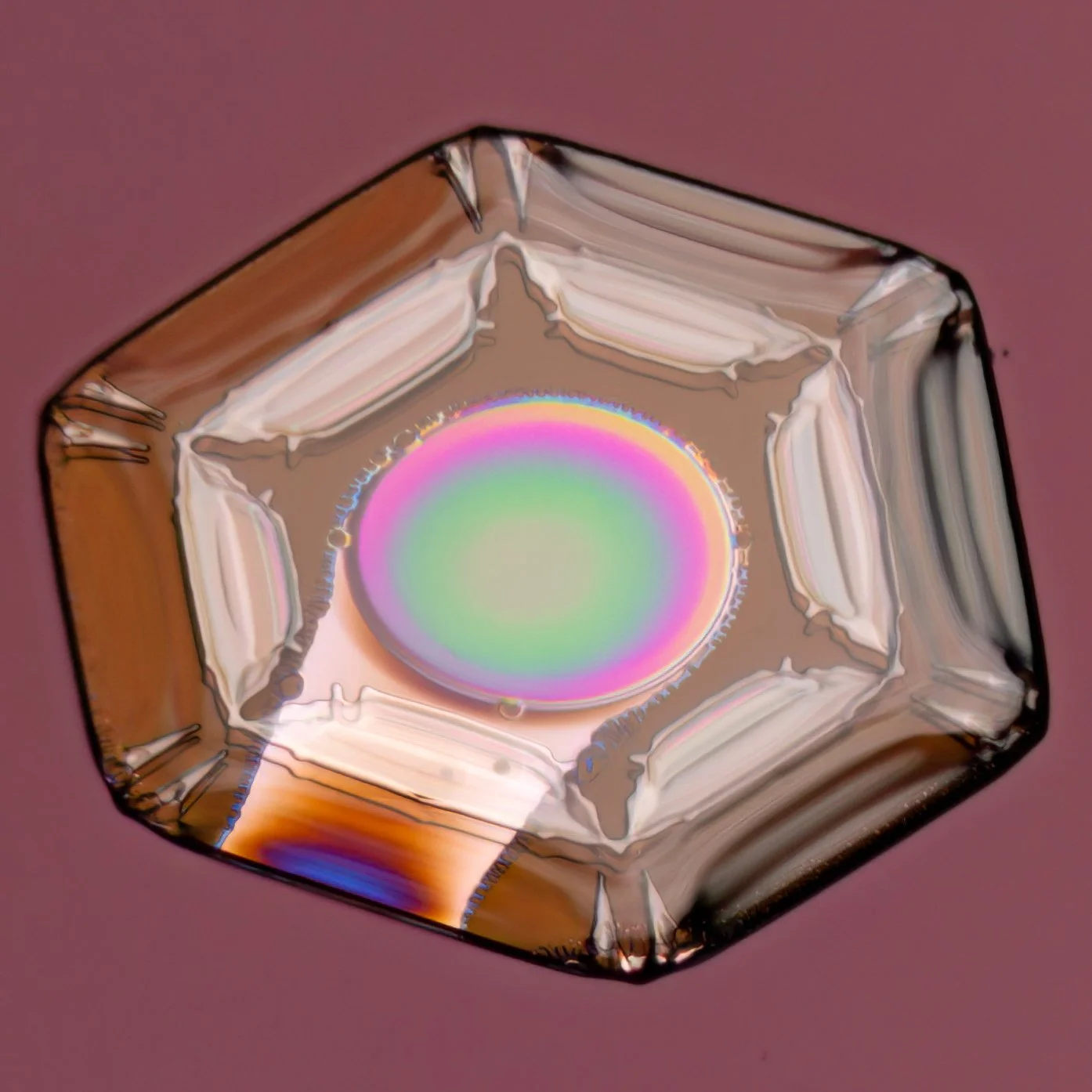 Macro photograph of a small, square-shaped crystal with a colorful, iridescent center, set against a pink background.