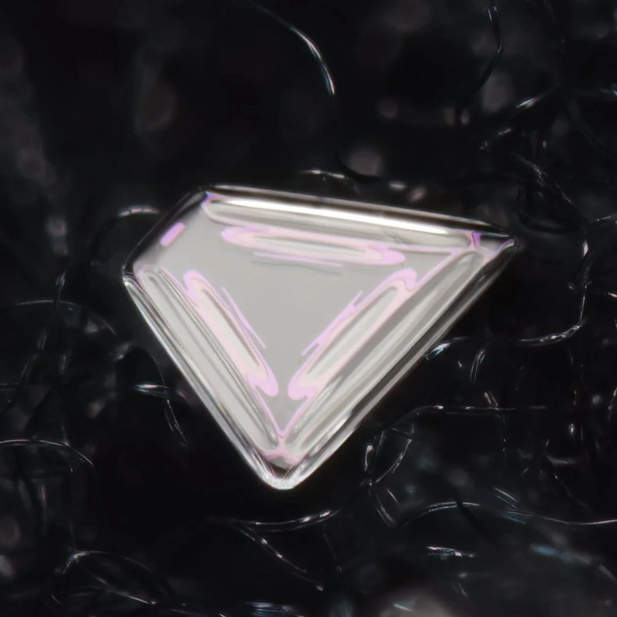 Macro photograph of a shiny, reflective diamond-shaped snow crystal with a pinkish hue, resting on a black textured surface.