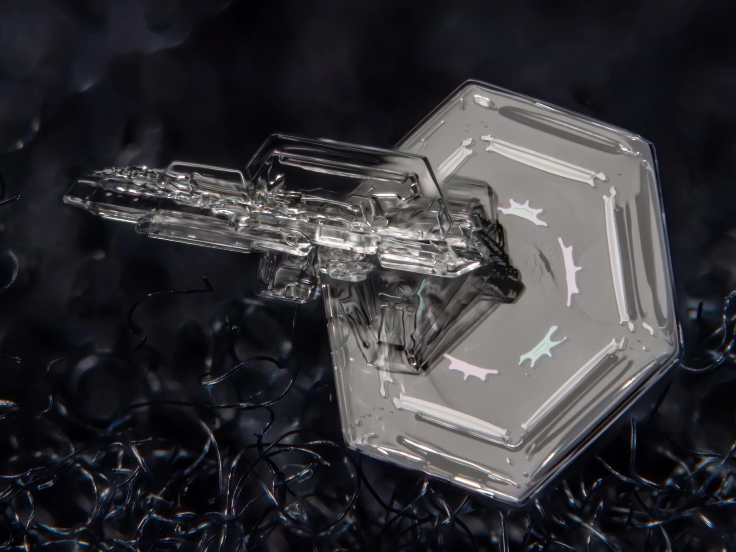 A macro photograph of two snow crystals attached to each other with the lower plate showing visible irridence.
