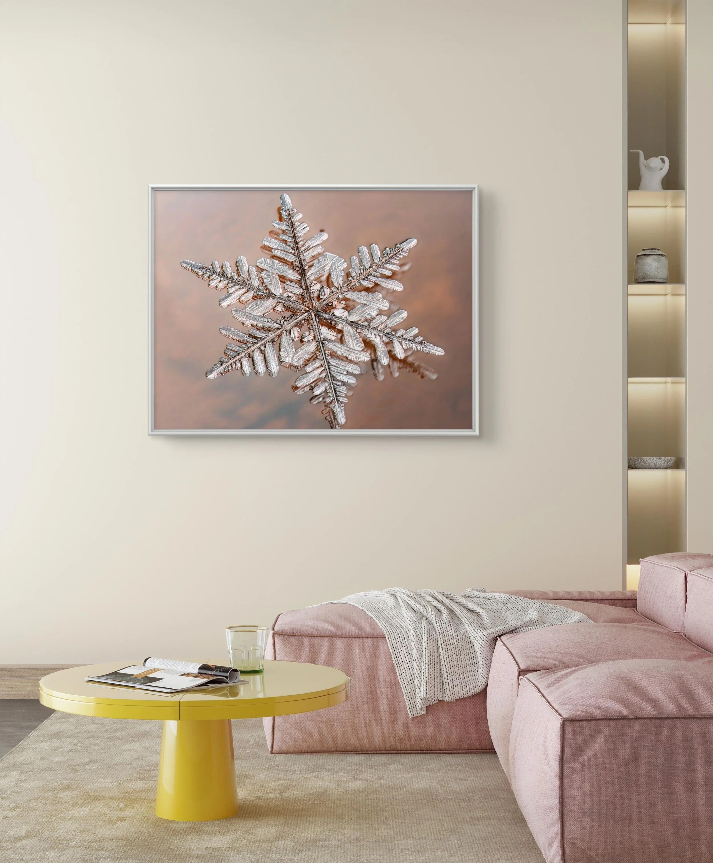 Living room with light-colored walls, a pink sectional sofa with a blanket, a yellow coffee table with a glass, magazines, and a remote, and a large framed picture of a snowflake hanging on the wall.