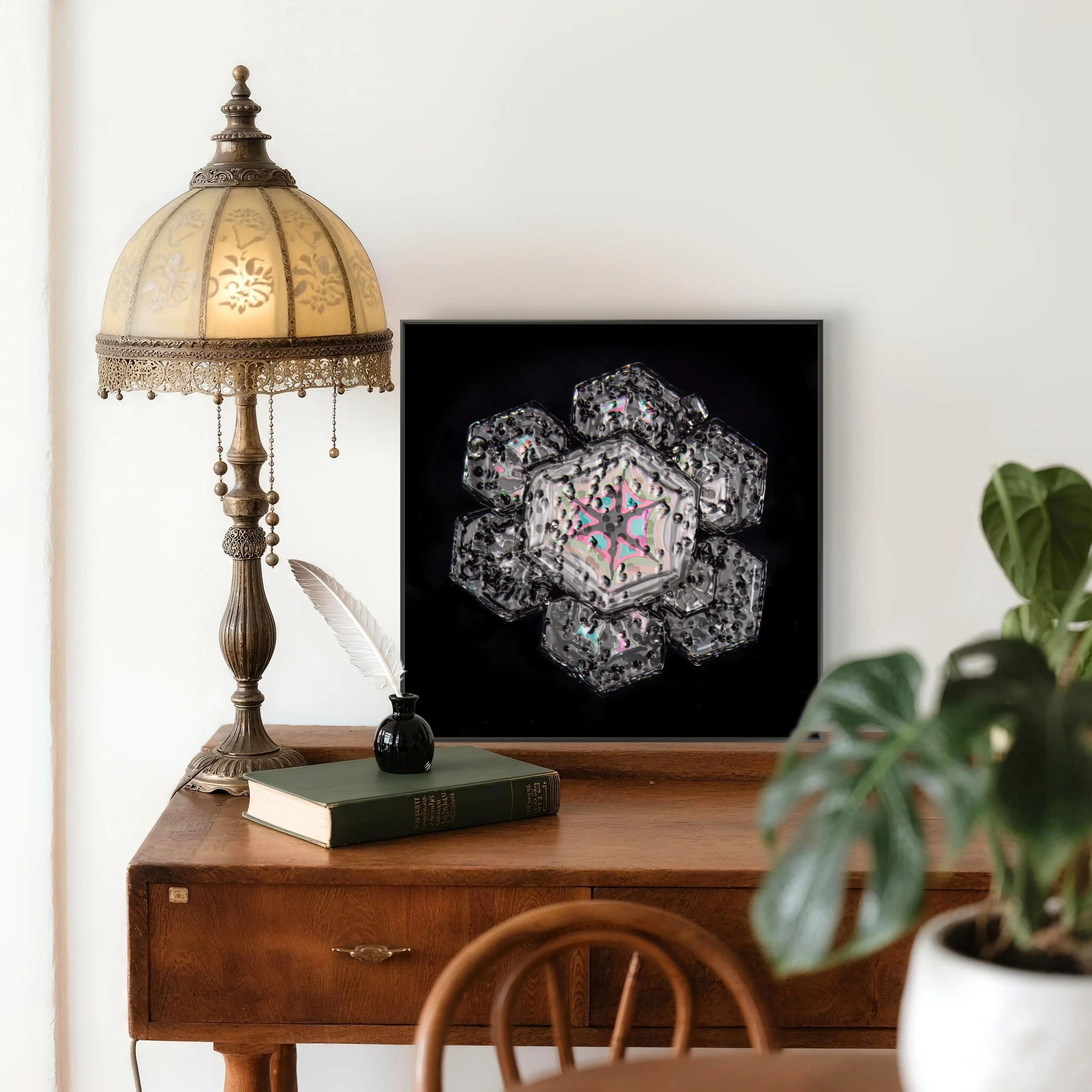 Decorative living room scene with a vintage table lamp, a framed geometric snow crystal with a colourful center structure, a closed book, a small black vase with a white feather, a potted plant, and a wooden chair in the foreground.