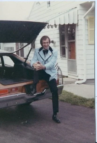 A man in a light blue jacket and black pants seated on the back of a brown car with an open trunk, smiling at the camera, outside a residential house with white siding and a small step.