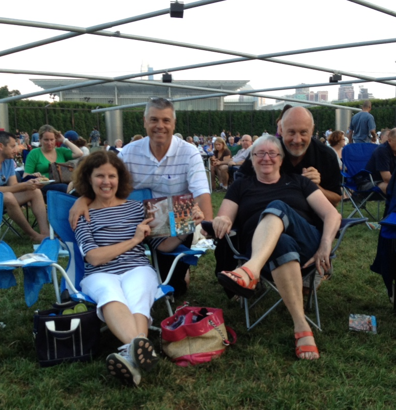 Four people sitting outdoors on lawn chairs at a gathering, with a crowd in the background, some holding plates, in an open park with structures overhead and a cityscape in the distance