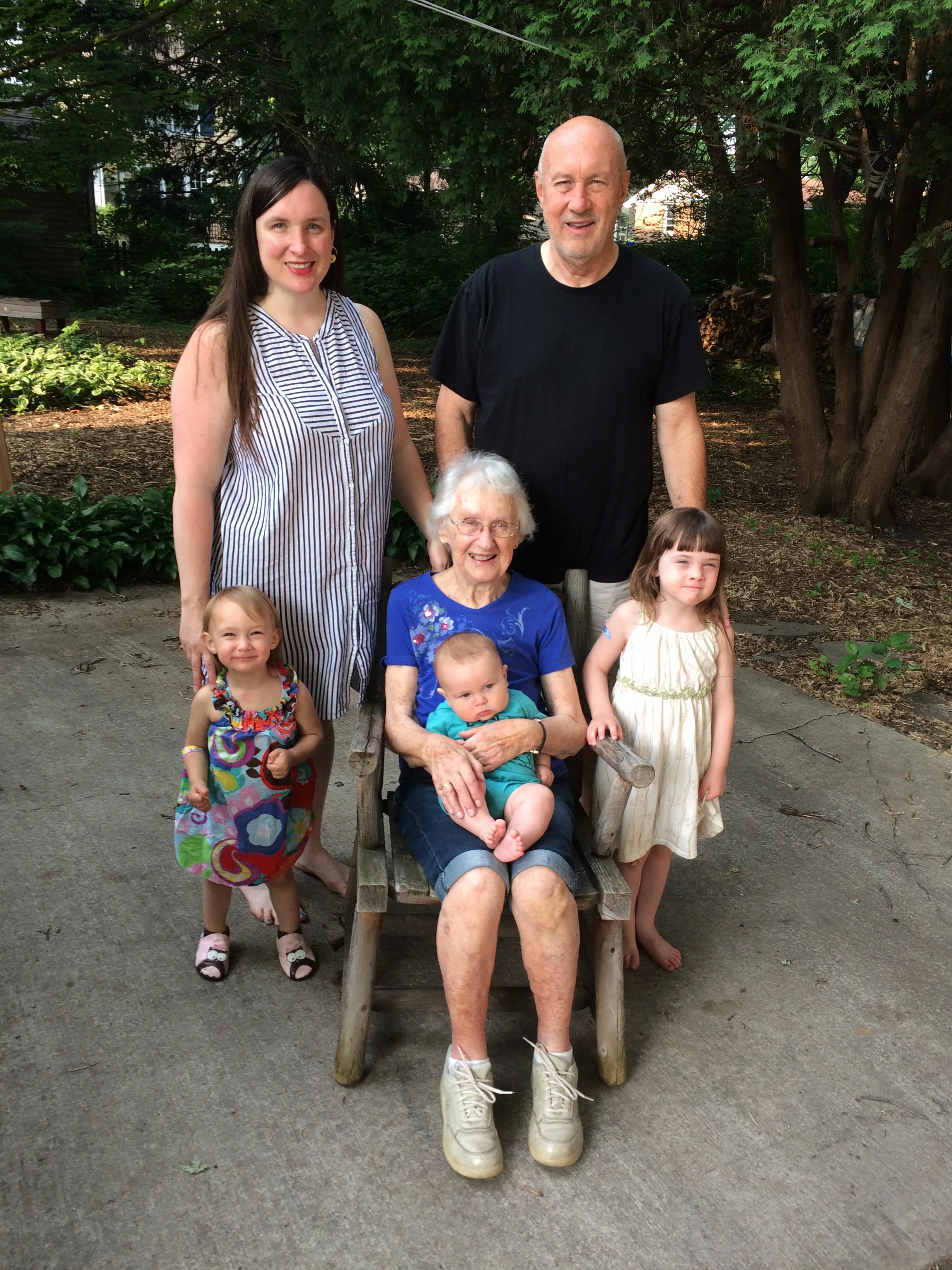 A multi-generational family photo taken outdoors featuring six individuals, including an elderly woman seated in a wooden chair holding a baby, a teenage girl, a young girl, an adult woman, and an adult man standing behind the elderly woman. They are surrounded by green trees and plants.