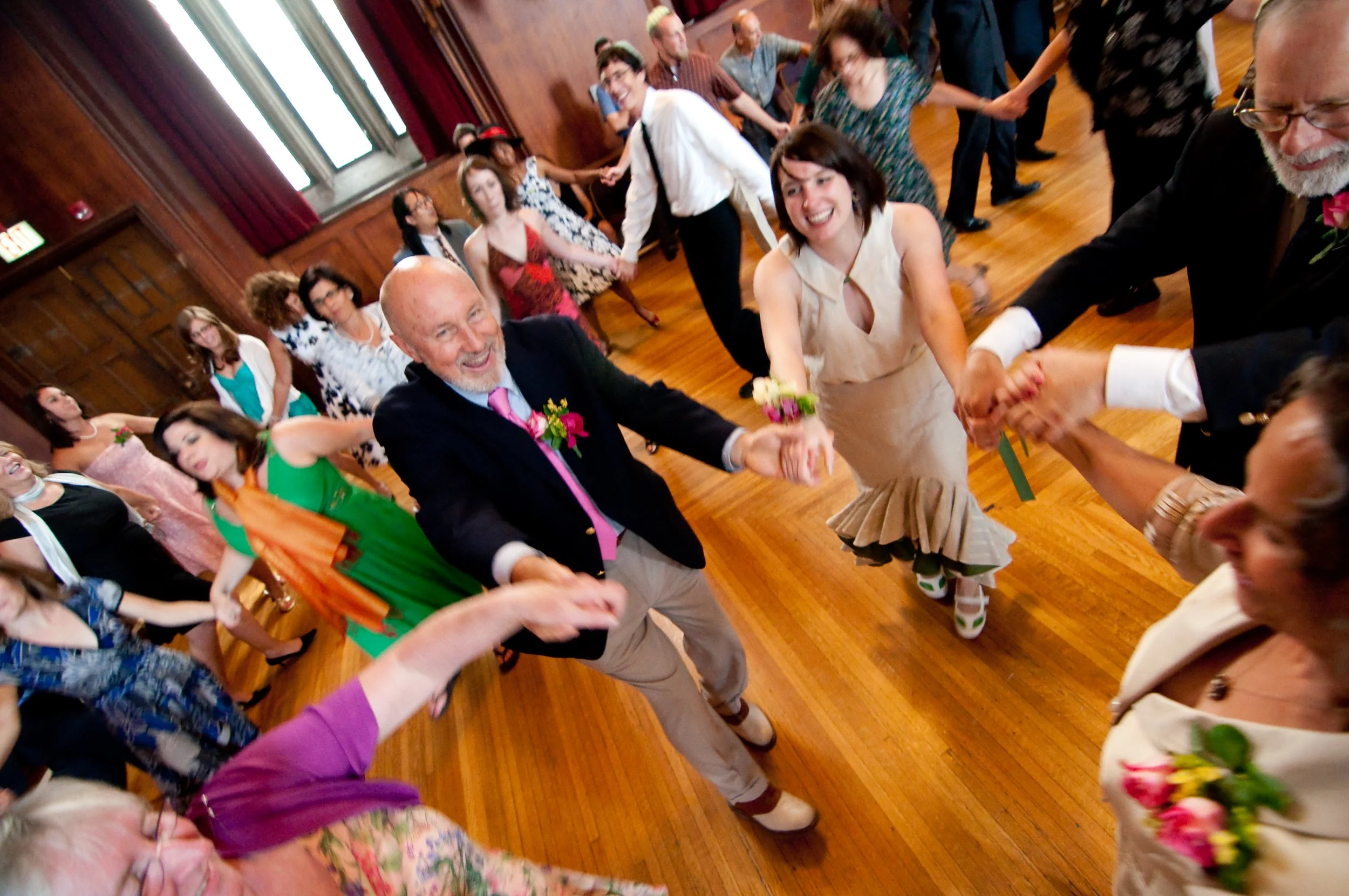 People dancing in a circle at a wedding reception, smiling and holding hands inside a wood-paneled hall.