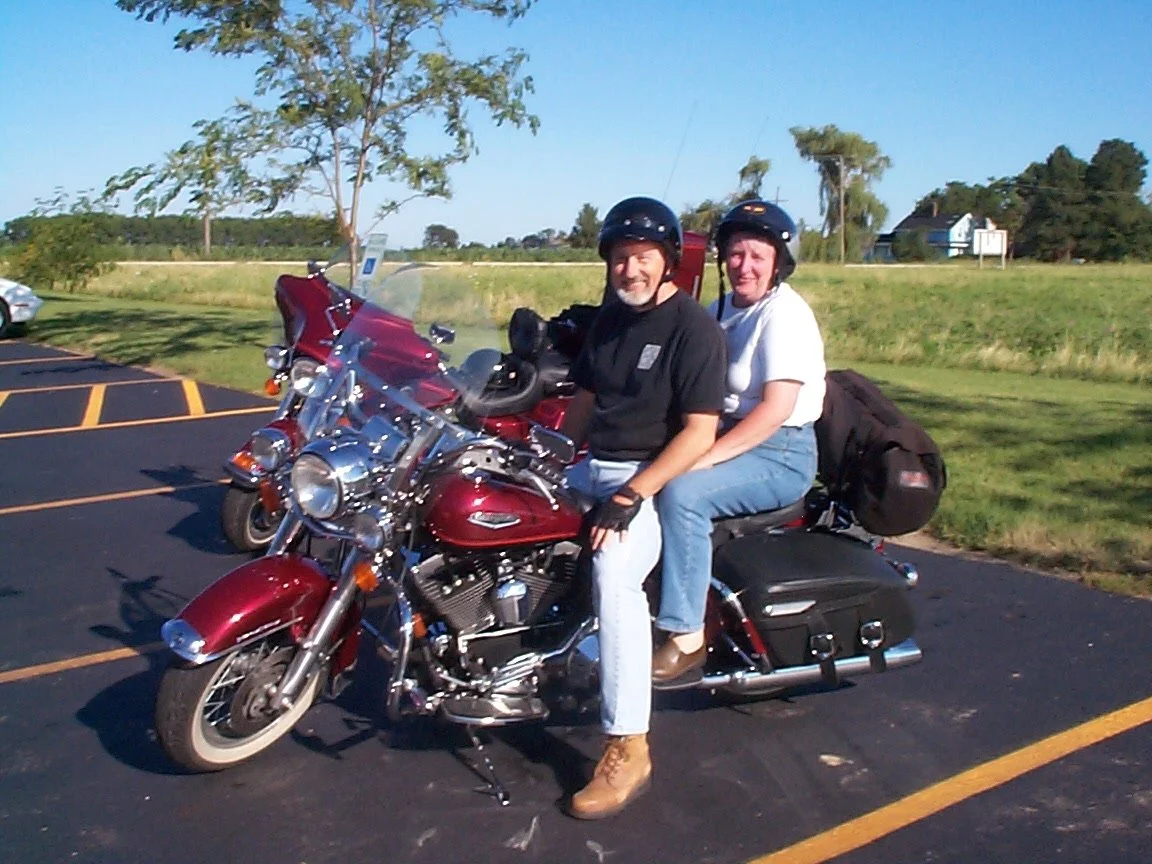 Two people sitting on a red touring motorcycle in a parking lot on a sunny day, wearing helmets.