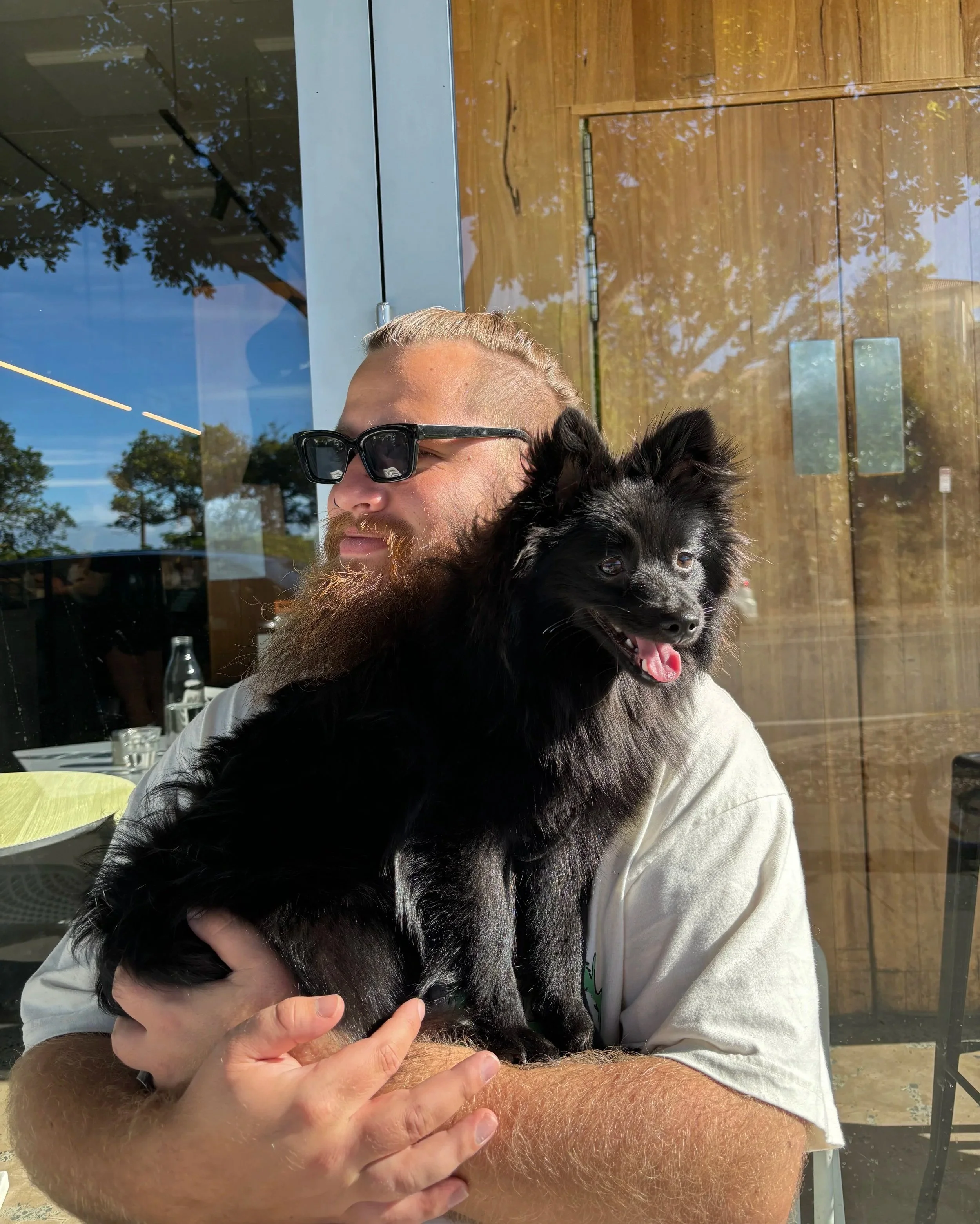 A man with blonde hair and a beard, wearing sunglasses and a white shirt, sitting inside a building and holding a small black dog with a thick coat and pointy ears. The man is smiling and the dog is looking happy with its tongue out.