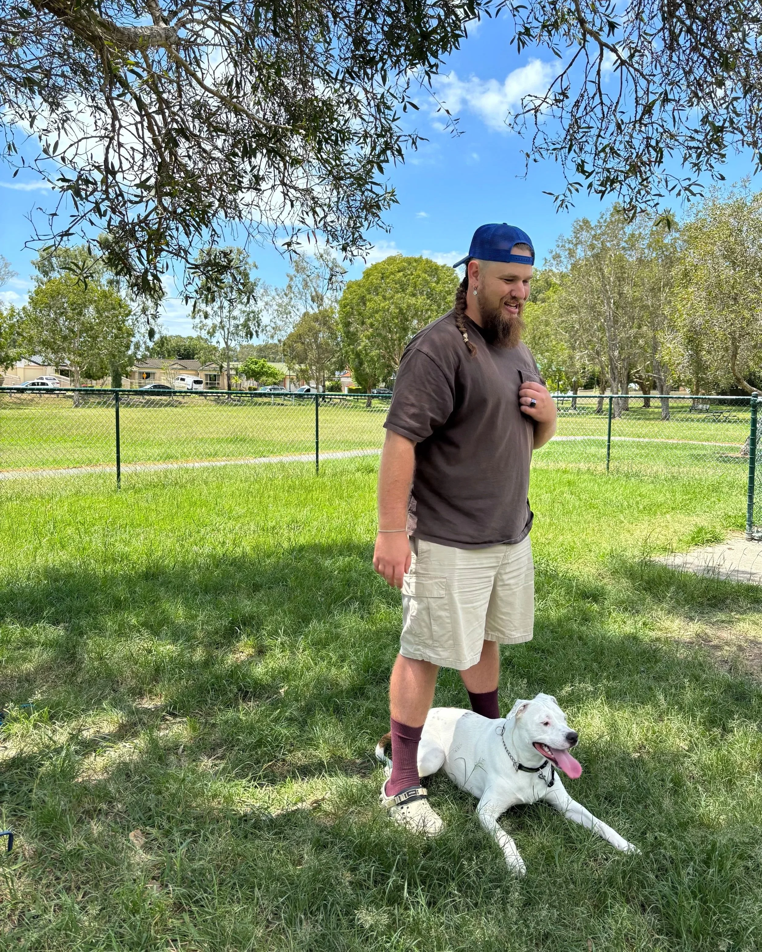A man with a beard, wearing a blue baseball cap, brown T-shirt, beige shorts, and maroon socks, standing on grass with a white dog lying down beside him. The man is smiling and holding his chest with one hand under a tree, in a park with a fence and 