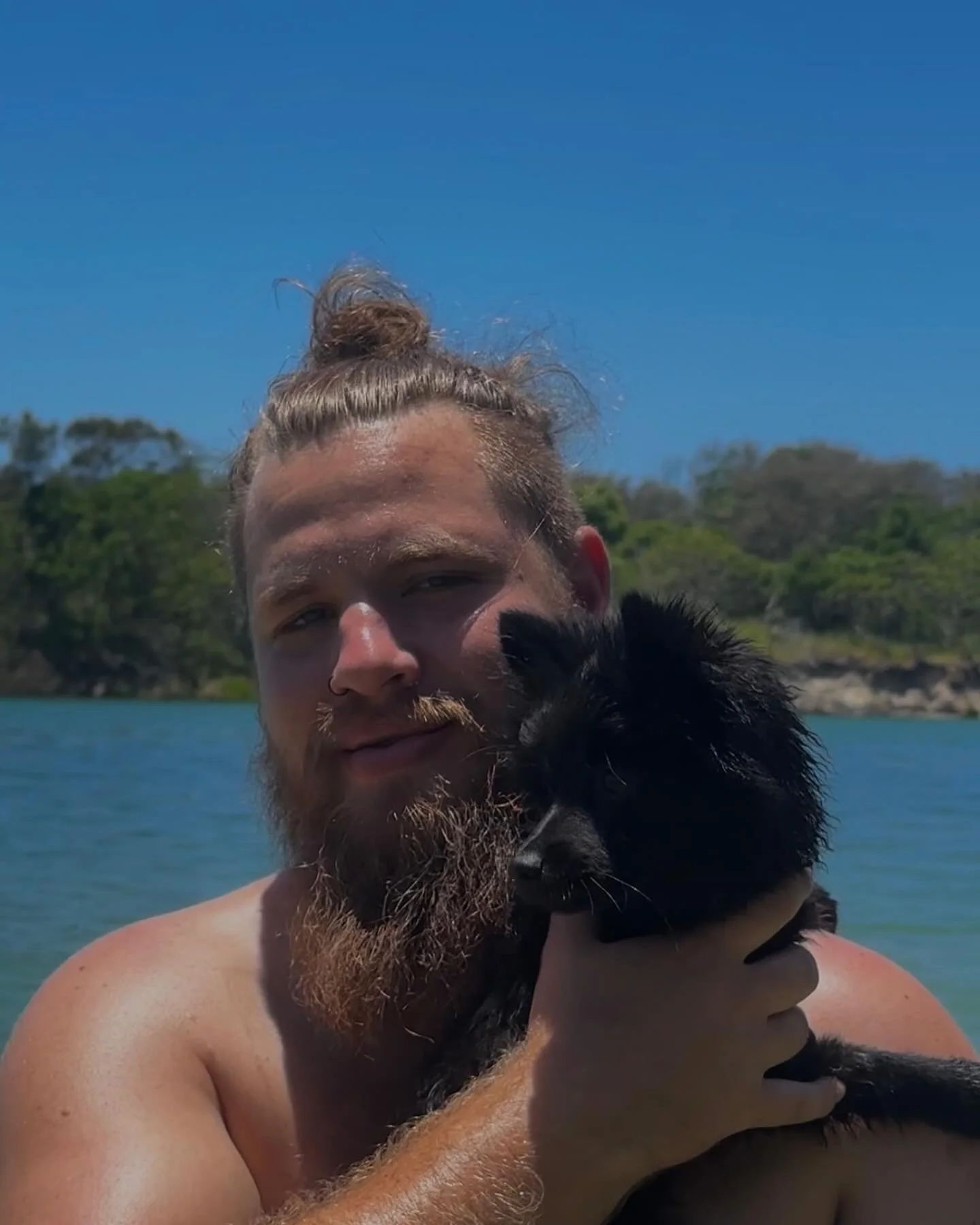 A man with a bun hairstyle and beard holding a black dog near water with a forested landscape and blue sky in the background.