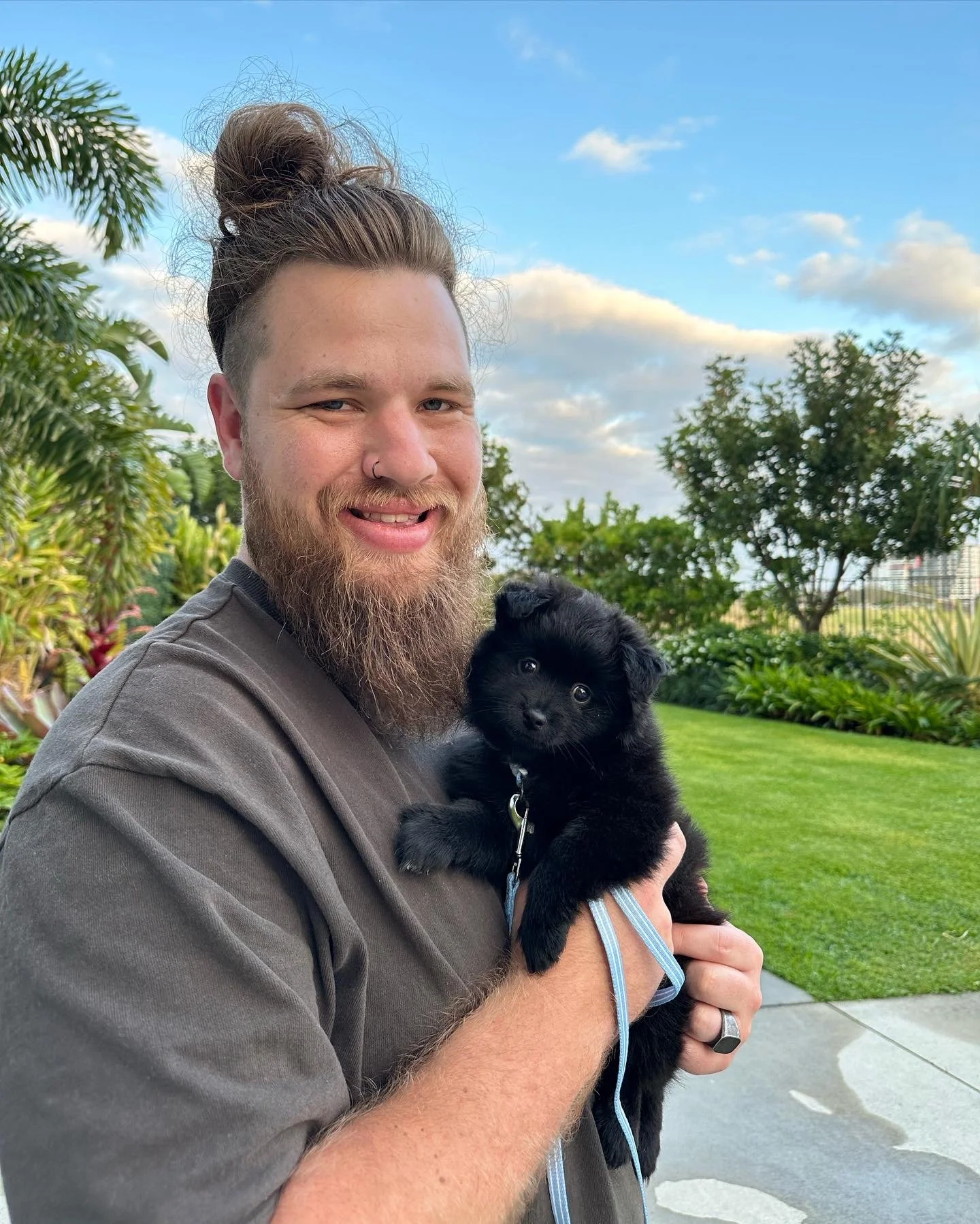 A man with a beard and bun hairstyle holding a black puppy outdoors in a garden with trees and grass, under a partly cloudy sky.
