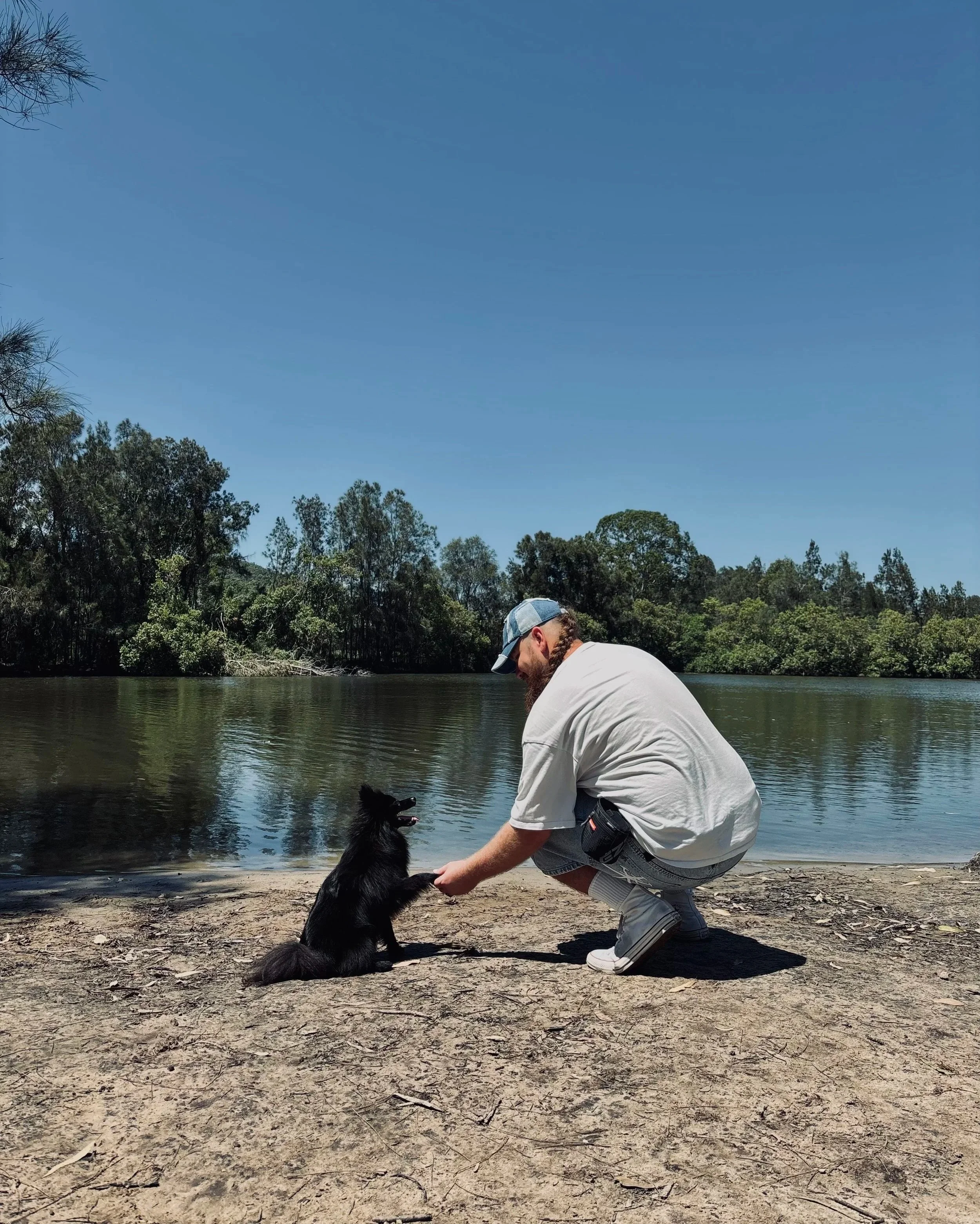 A man crouching on the beach near a river, holding the paw of a black dog sitting in front of him. The scene includes trees in the background under a clear blue sky.