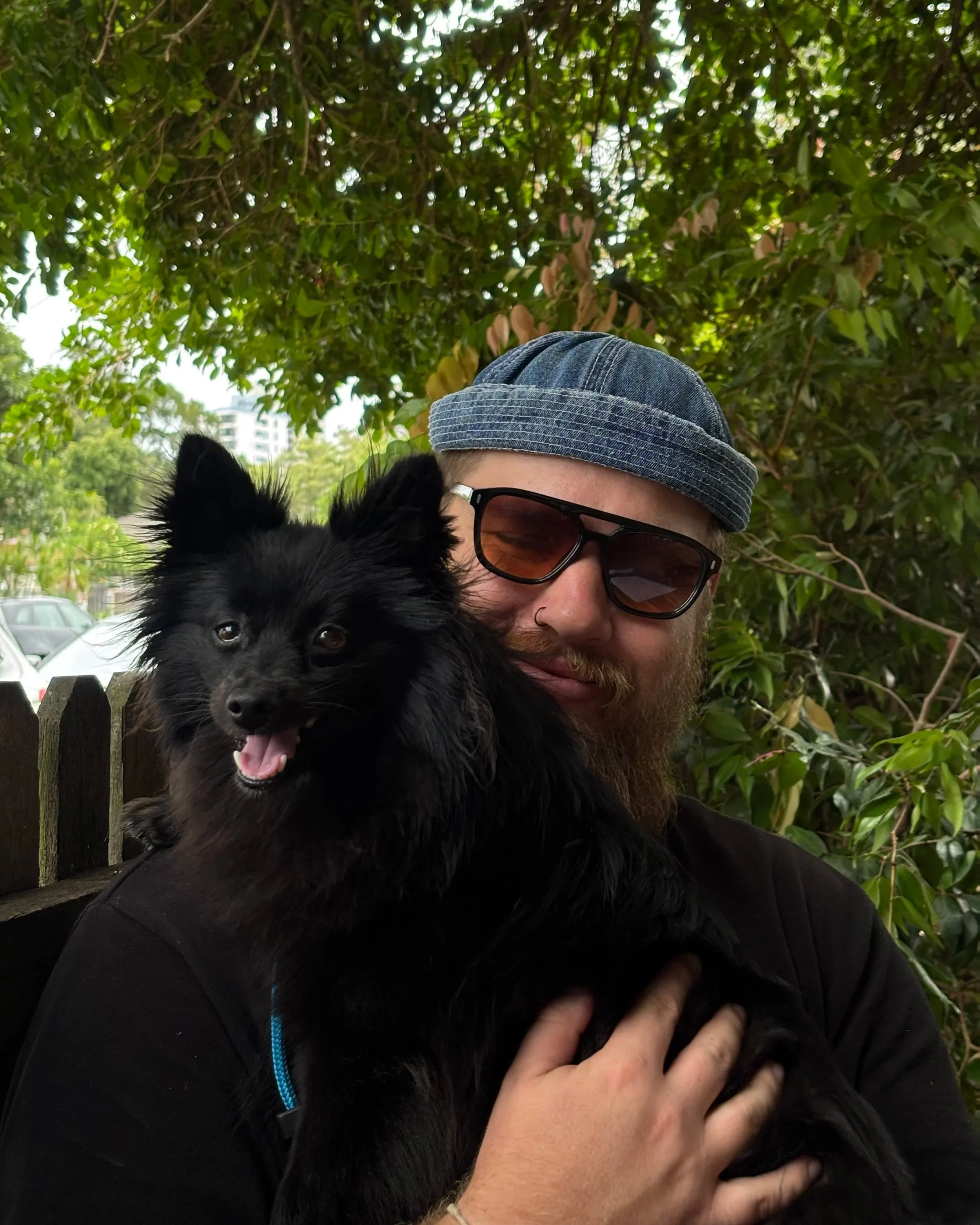 A man with a beard, sunglasses, and a nose ring, is holding a black fluffy dog with pointy ears in a green outdoor setting.