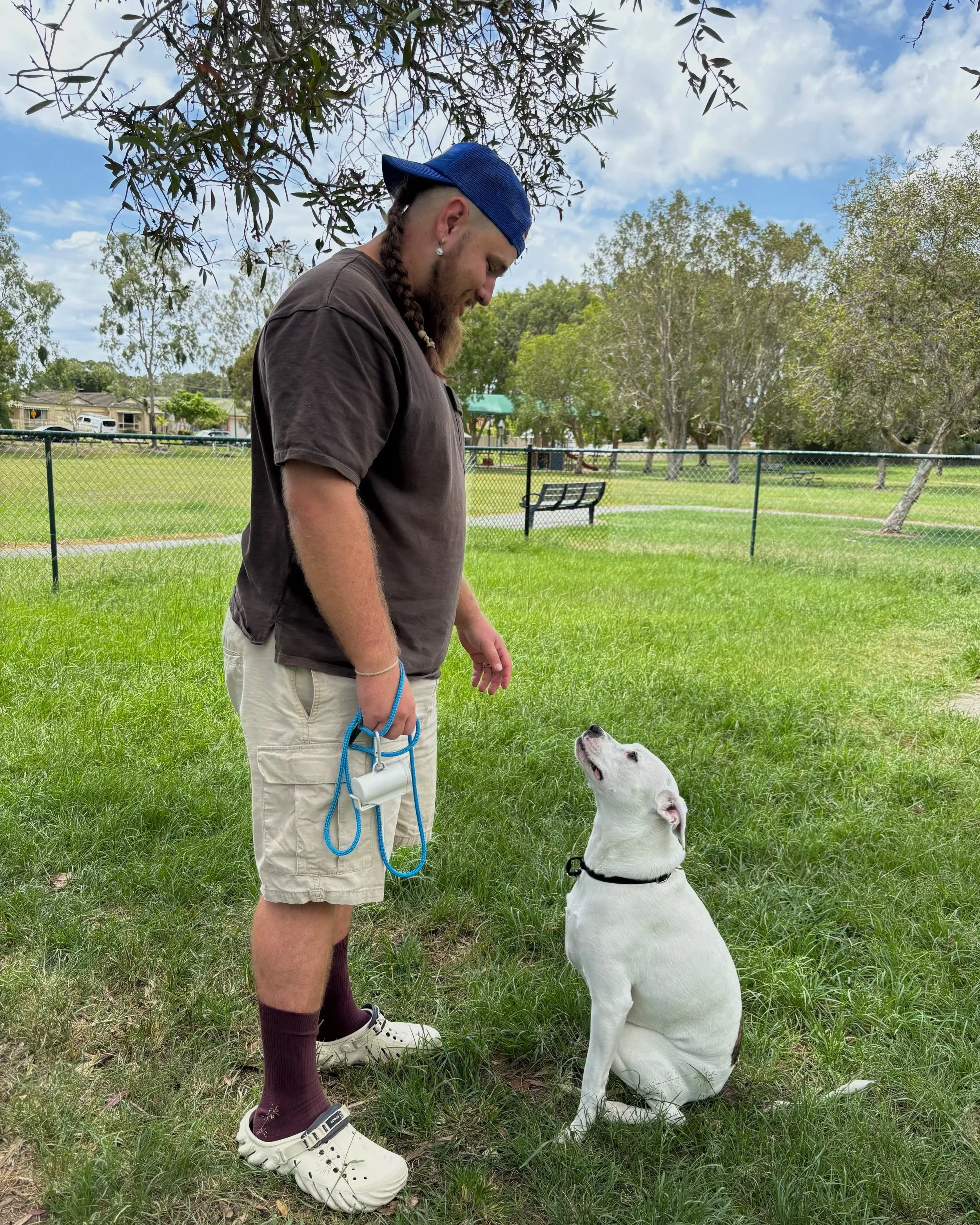 A man with long braided hair, wearing a blue cap, a brown t-shirt, khaki shorts, and white sneakers, standing on grass with a leash in his hand, looking down at a white dog with black spots sitting attentively, looking up at him, in a park with trees