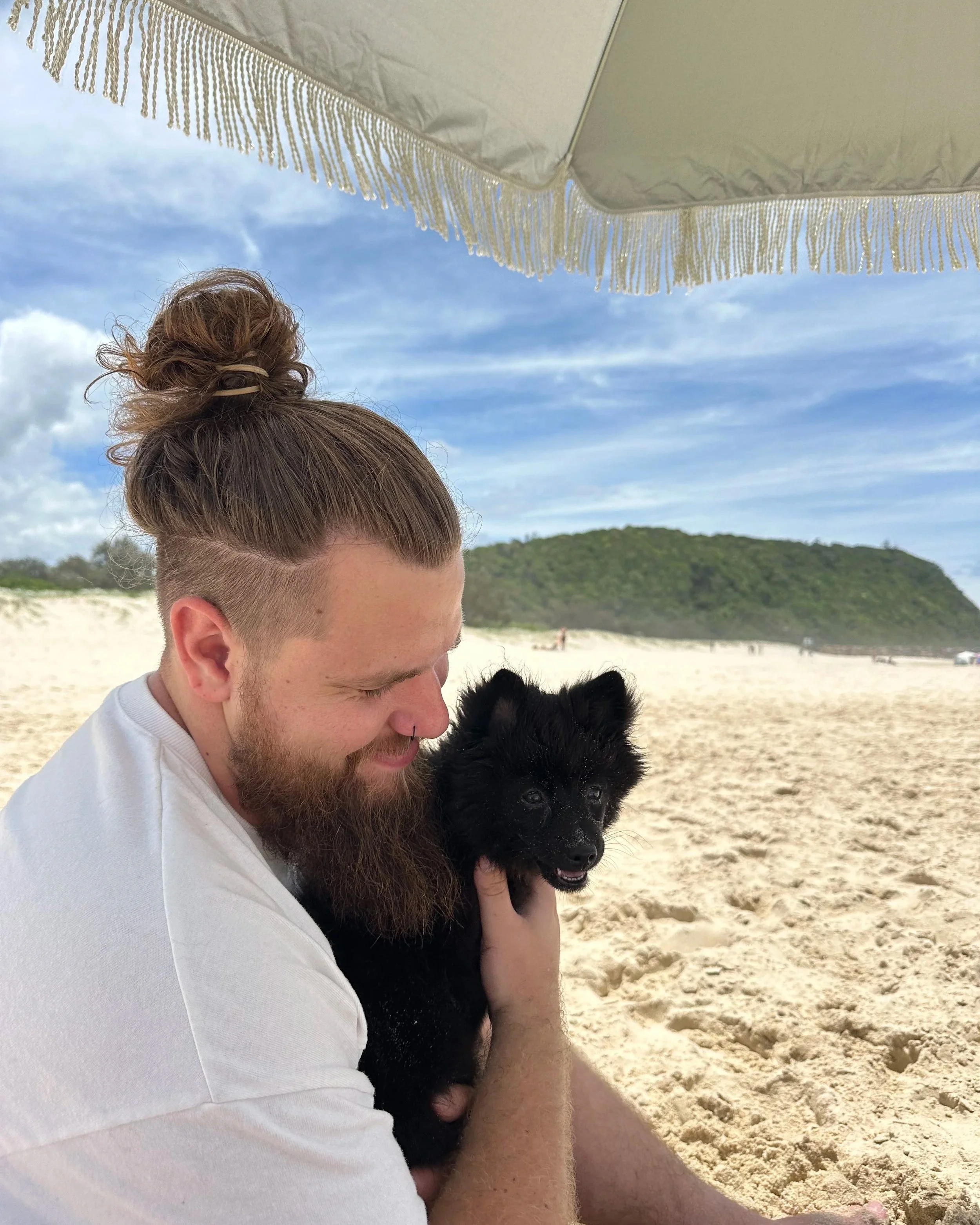 A man with a bun hairstyle and a beard is holding a small black puppy at the beach under a white canopy. The puppy has sand on its face and looks happy. The background shows sandy beach, green hills, and a blue sky with some clouds.