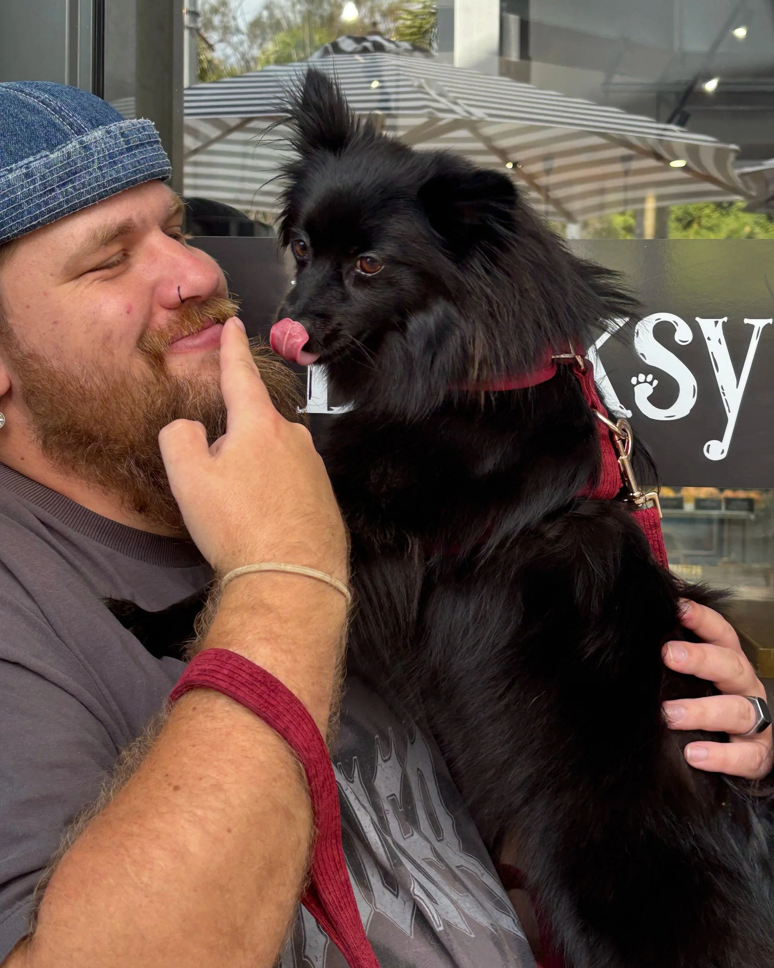 A man with a beard and hat smiling as a black dog licks his nose, sitting on his chest. They are outside near a window with a striped umbrella in the background.