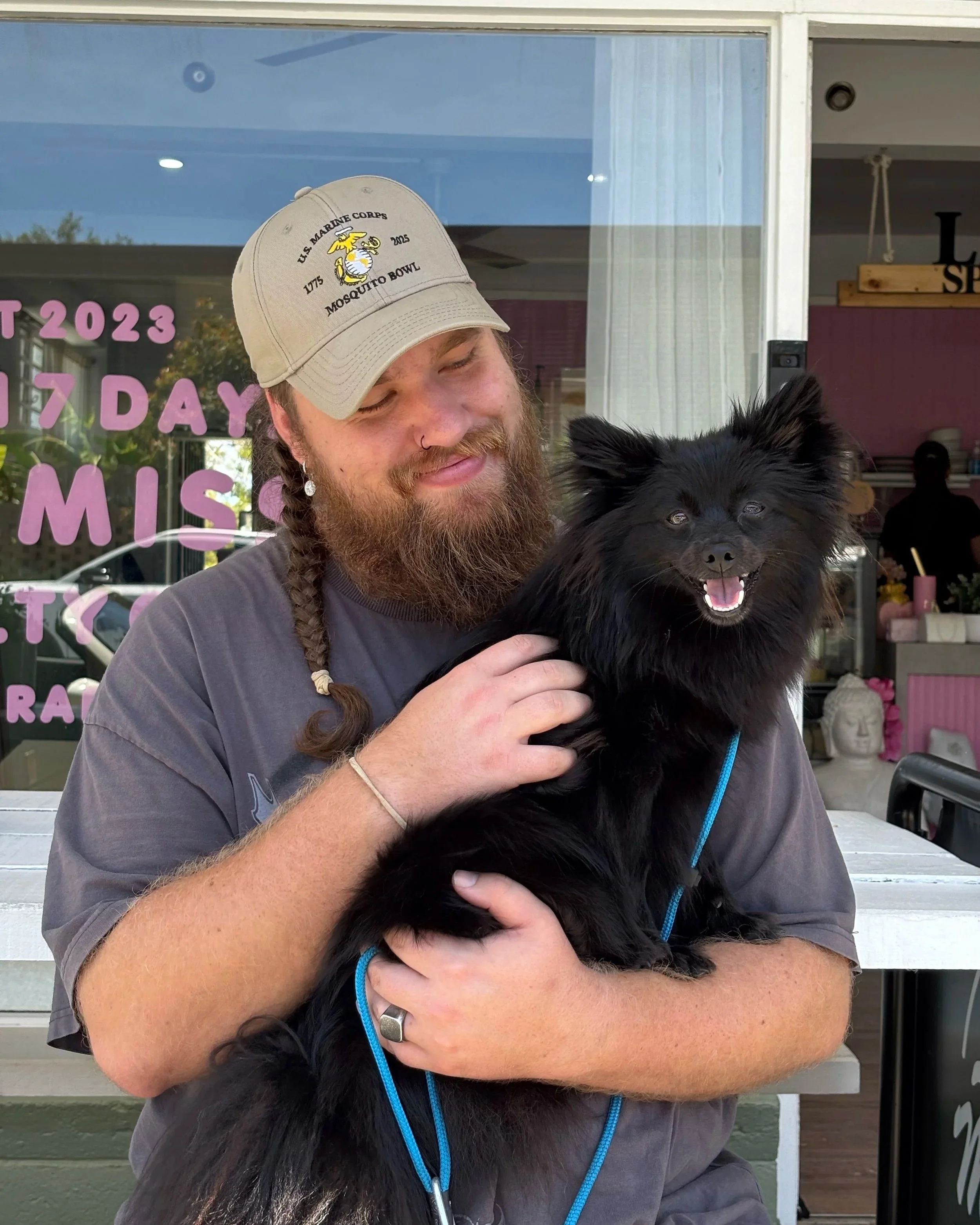 A man with a beard and braided hair, wearing a beige baseball cap and a gray t-shirt, holding a smiling black dog with fluffy fur in front of a storefront window.