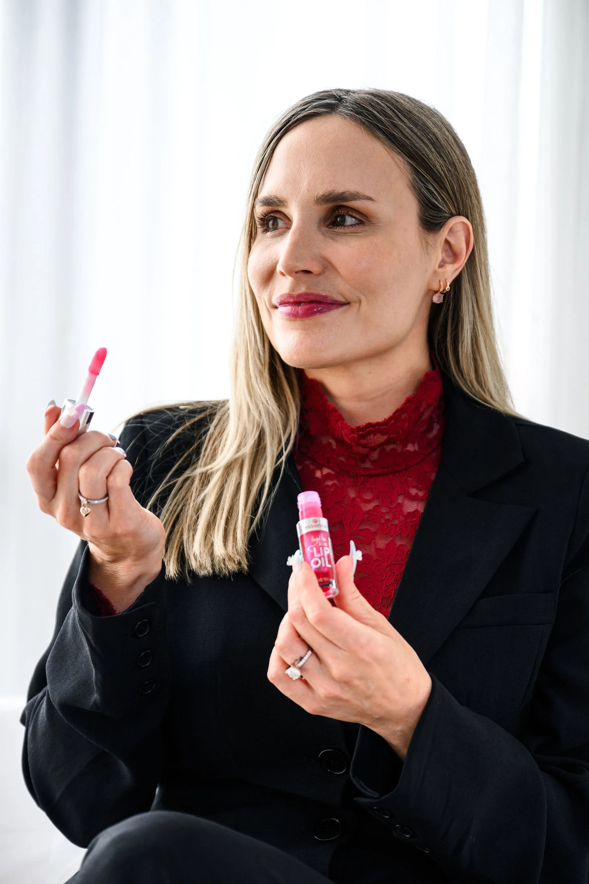 A woman holding a lip gloss and lip oil, looking to the side with a slight smile, dressed in a black blazer and a red lace top, sitting indoors near white curtains.
