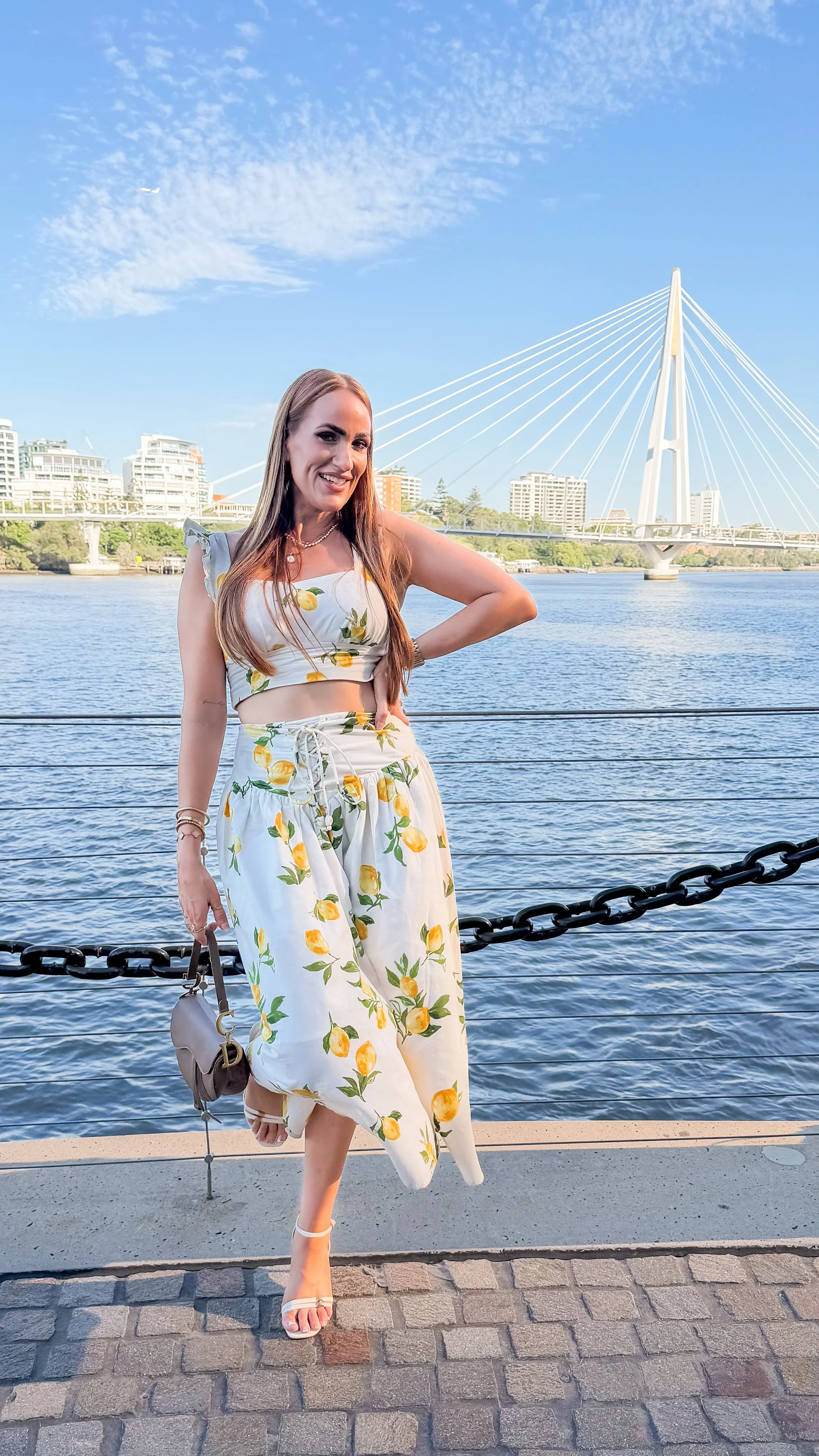 A woman standing by a waterfront with a cityscape and bridge in the background, wearing a lemon-printed outfit and white sandals.