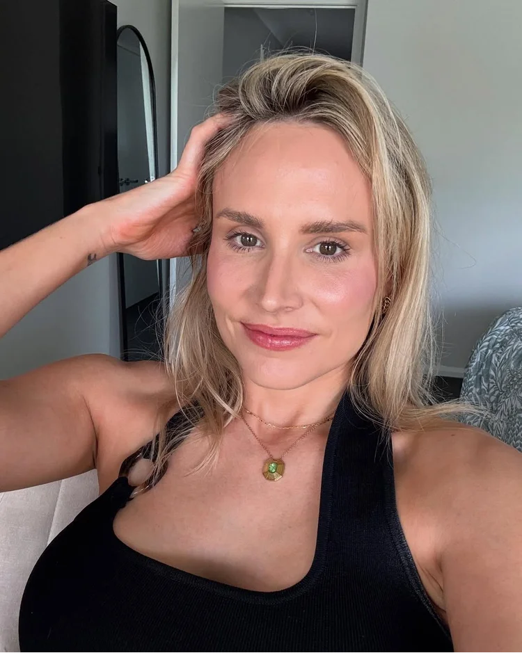 A woman with blonde hair wearing a black tank top and a gold necklace, smiling and holding her hand to her head, sitting on a couch in a living room.