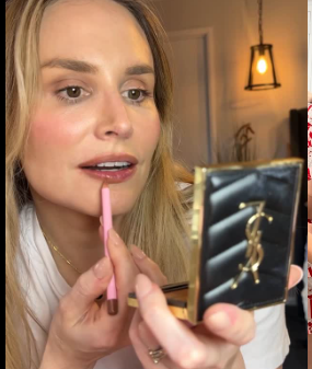 A woman applying makeup while looking into a mirror in a warmly lit room.