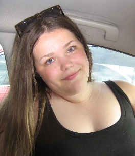 Young woman with long brown hair, wearing a black tank top and sunglasses on her head, smiling inside a car.