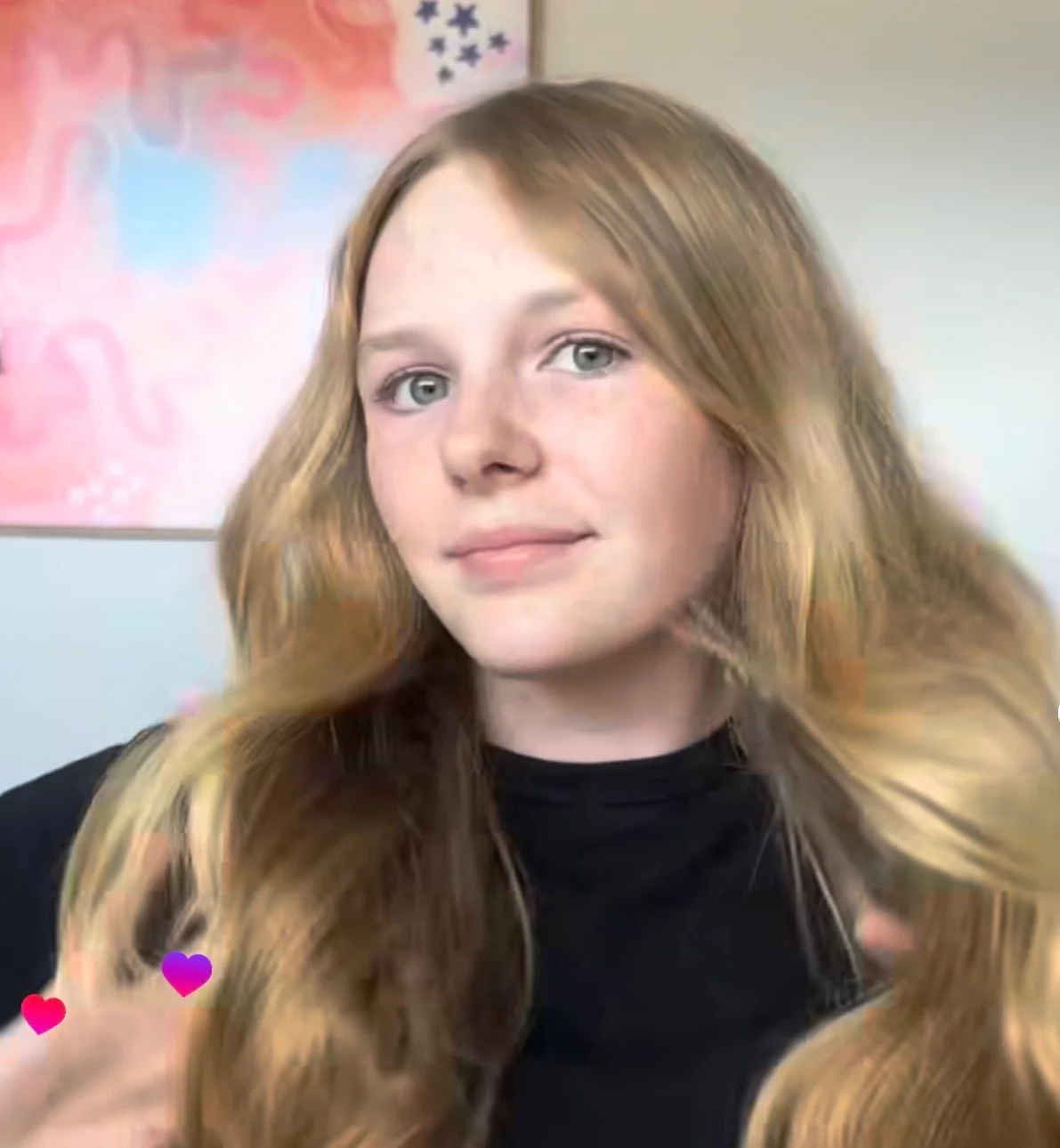 A young woman with long, wavy red hair and blue eyes, wearing a black top, is sitting indoors against a colorful background with pink, blue, and black artwork.
