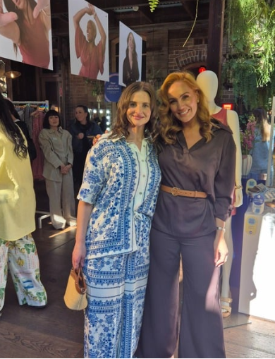 Two women posing together in a store or gallery, with clothing displays, mannequins, and hanging photographs in the background.