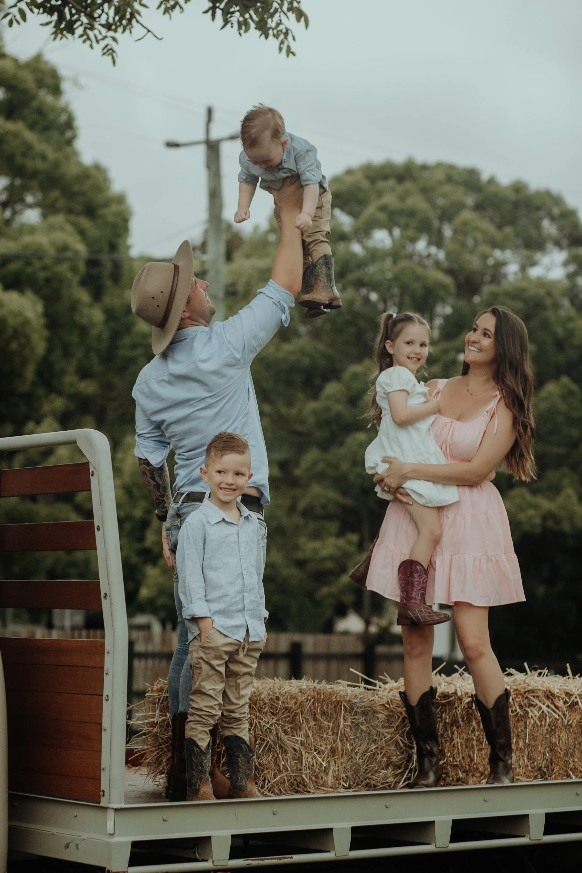A man with a cowboy hat lifts a young boy in the air at a farm, while two children and a woman stand nearby on a hay-covered float, all dressed in country-style clothing, with trees and power lines in the background.