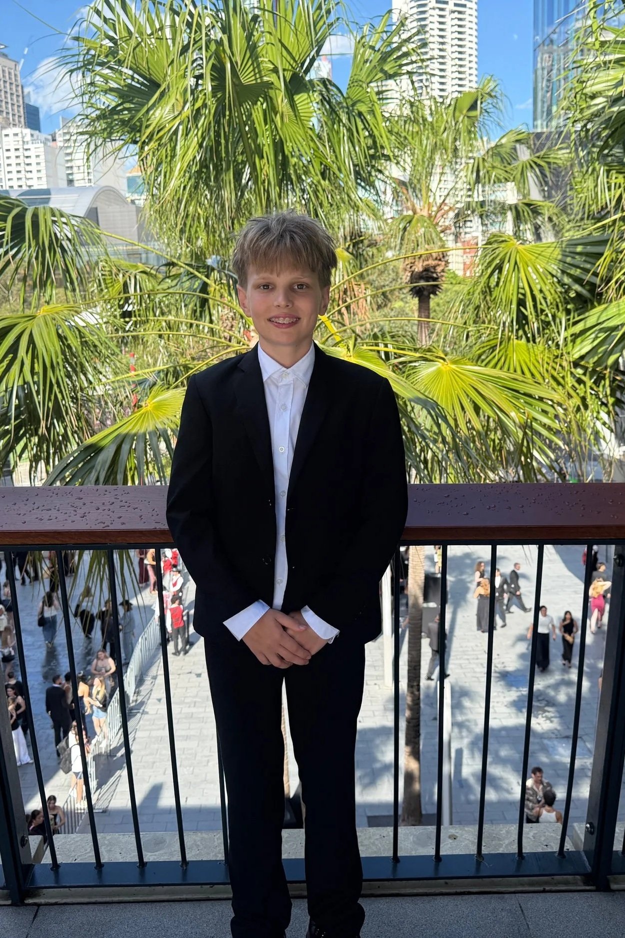 Young boy in a black suit jacket and white shirt standing in front of a railing, with urban buildings and palm trees in the background.