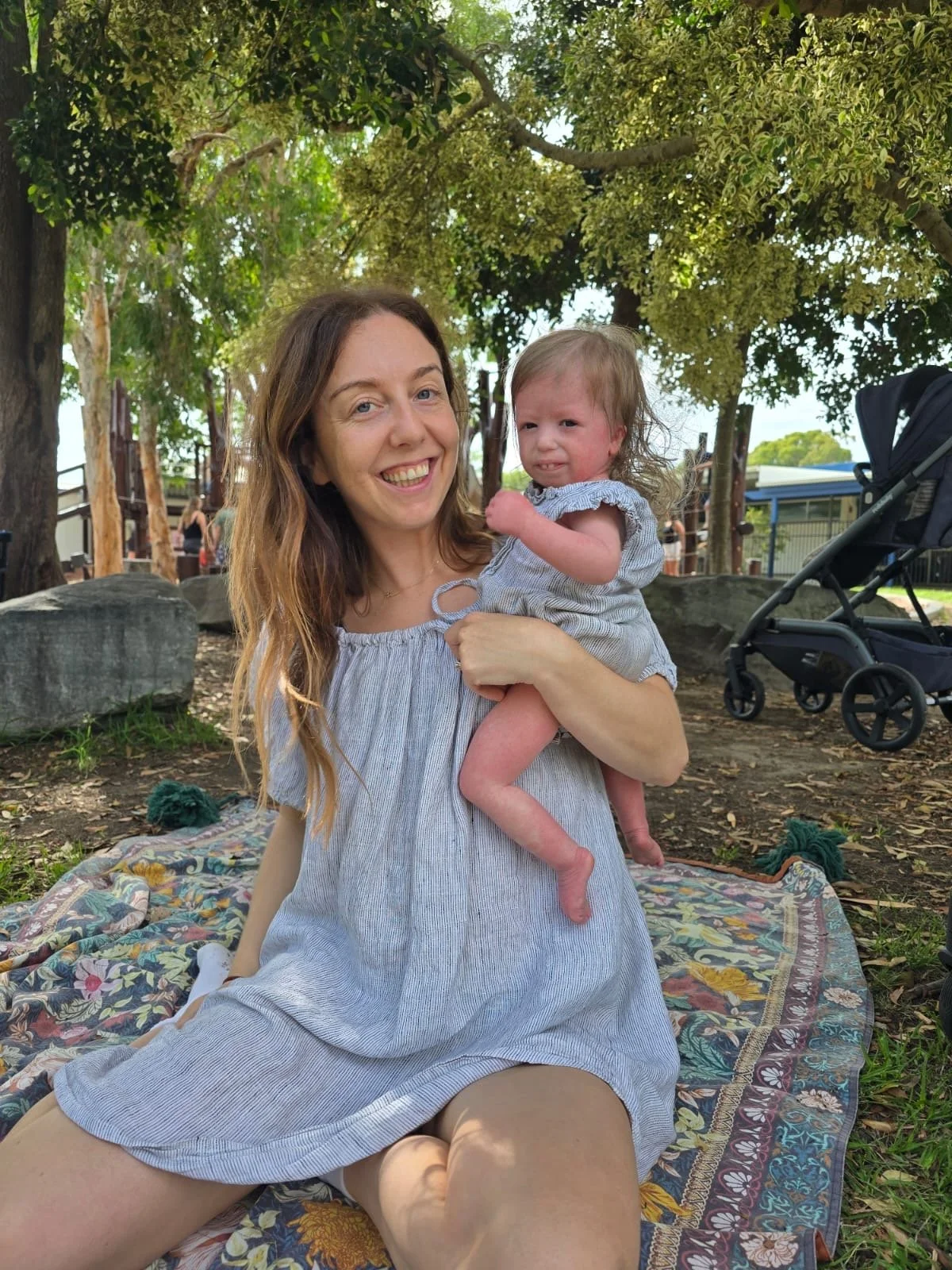 A woman smiling and holding a young girl on her lap outdoors in a park with trees, rocks, and a stroller nearby.