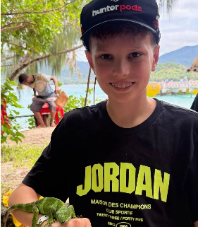 Young boy wearing a black cap and black T-shirt with bright yellow text, smiling outdoors near water with trees and mountains in the background, holding a turtle.
