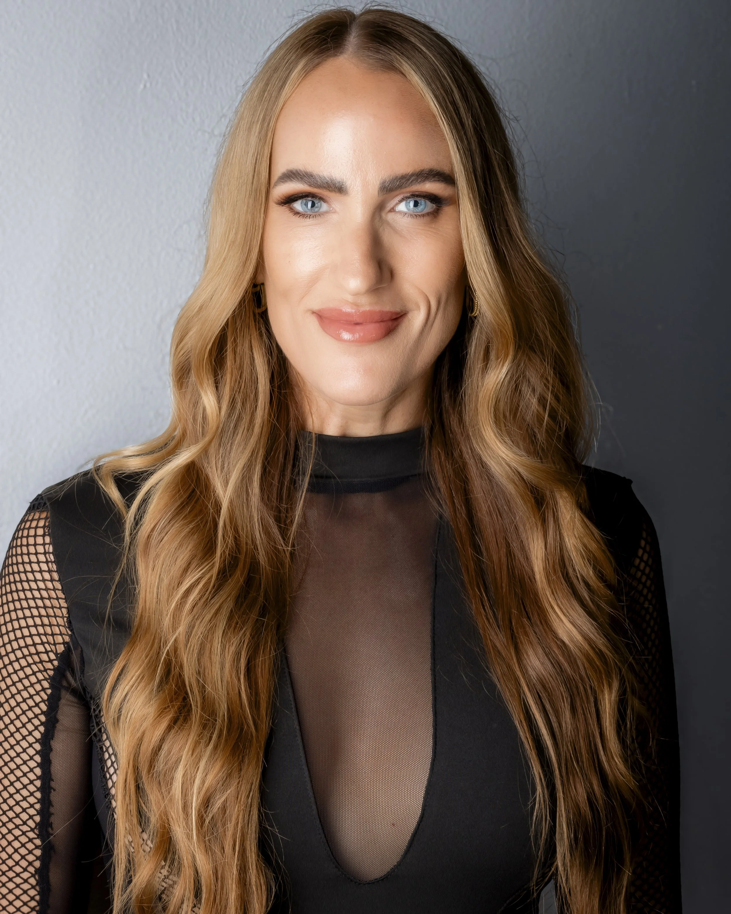 A woman with long, wavy blonde hair and bright blue eyes smiling, wearing eye makeup, coral lipstick, black mesh top, and hoop earrings, standing against a textured gray background.