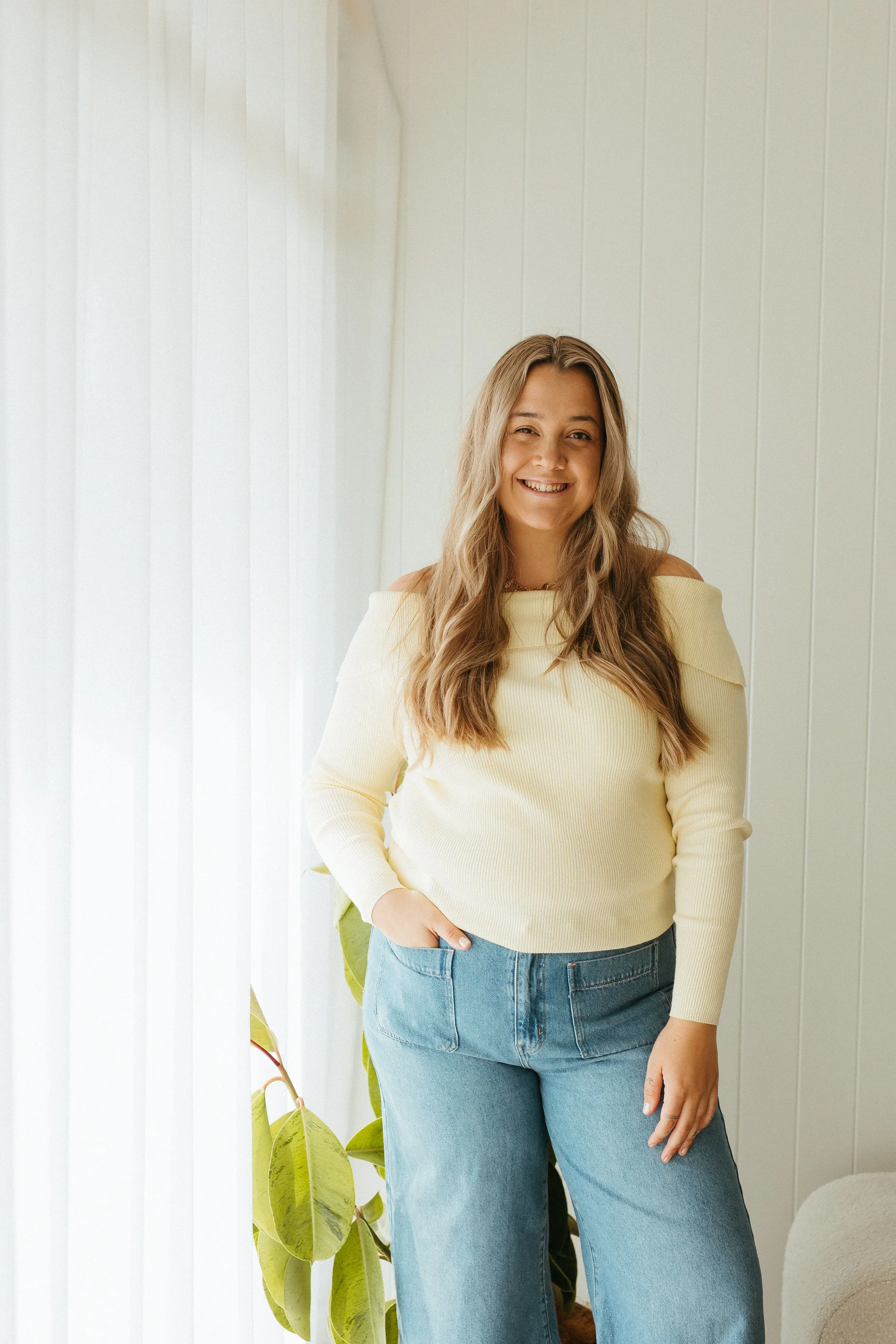UGC and content creator  A woman with long wavy blonde hair smiling and standing indoors near a window with sheer white curtains, wearing a light yellow sweater and blue jeans, with a large green plant behind her.
