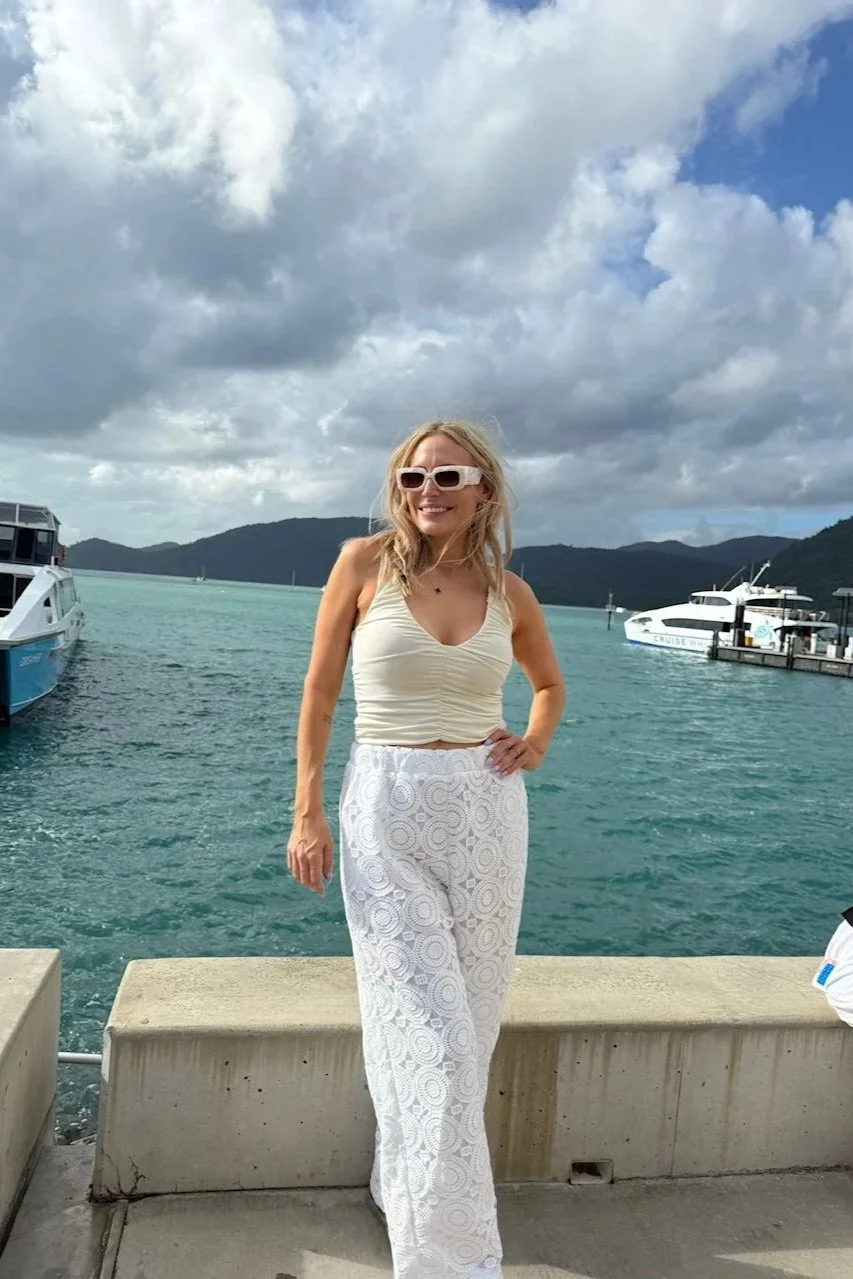 A woman with blonde hair, wearing white sunglasses, a beige sleeveless top, and white lace pants, stands on a concrete dock near the water with boats and mountains in the background.