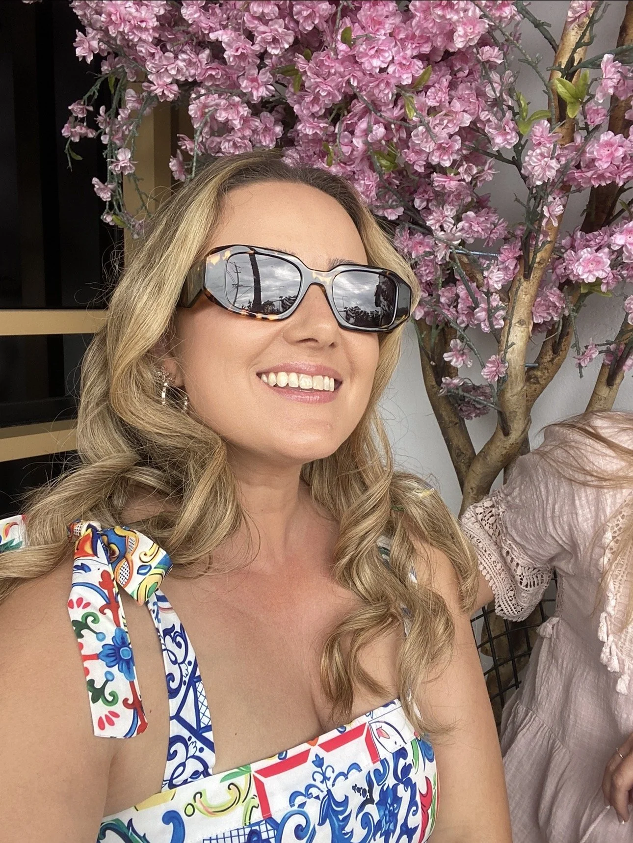 A smiling woman with long blond hair wearing sunglasses and a colorful dress with bow shoulder ties, standing in front of a pink flowering tree.