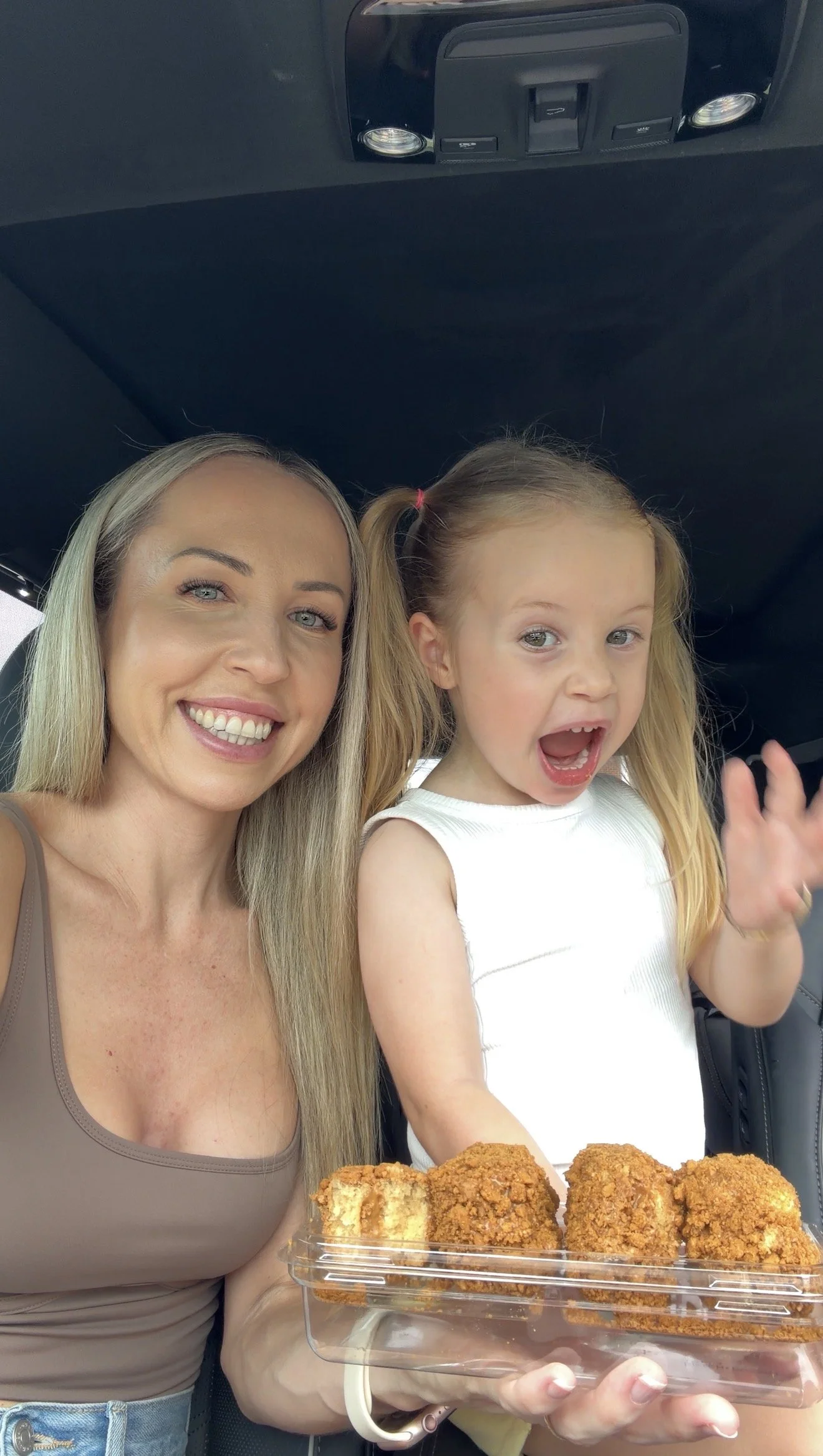 A smiling woman with blonde hair and a young girl with red hair, both in a car, holding a container of four fried chicken pieces or similar food items.