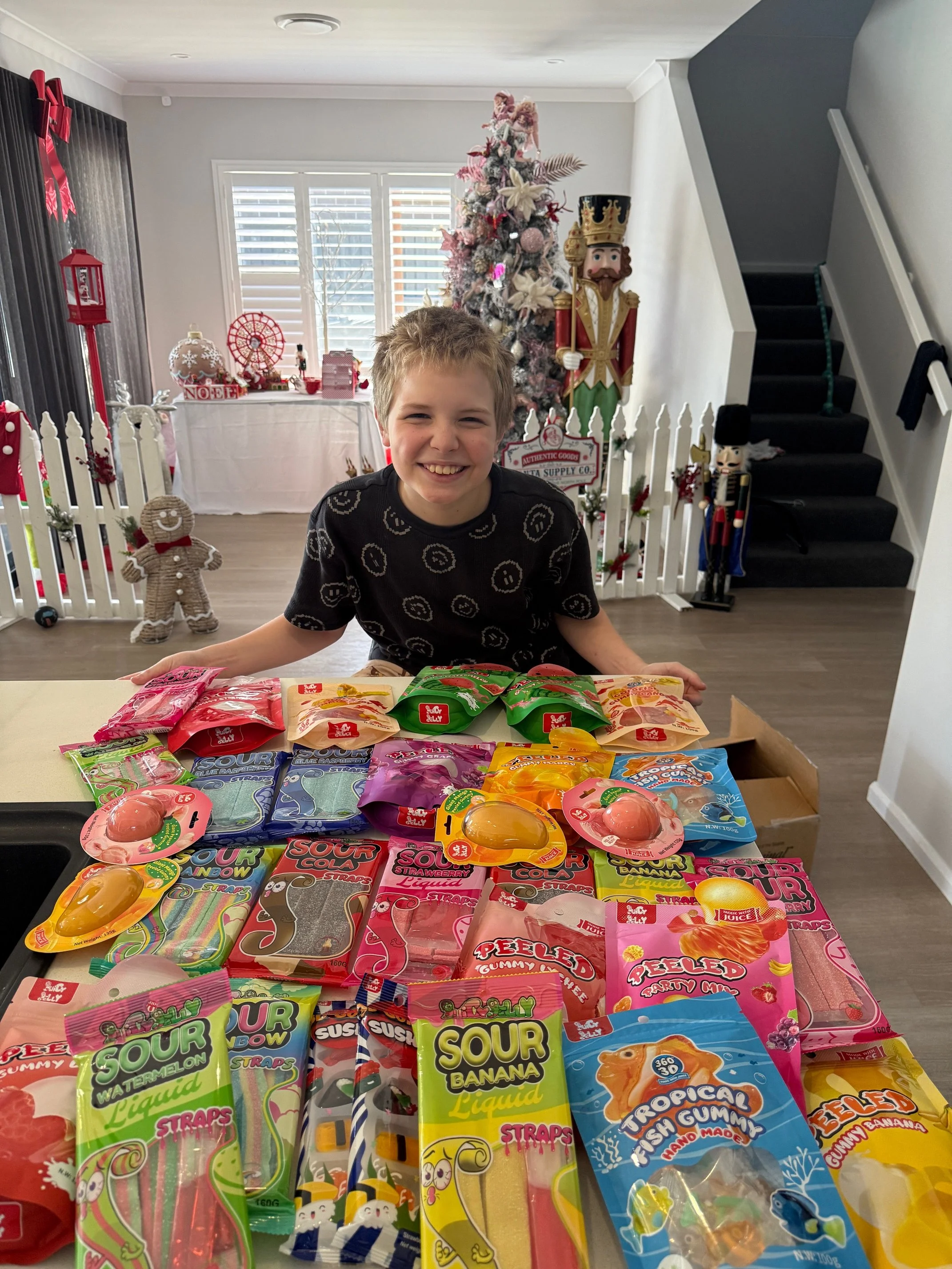 A smiling boy in a black t-shirt with white swirls sitting at a table with various colorful fruit-flavored candy products, with Christmas decorations including a Christmas tree, nutcracker, gingerbread man, and other holiday ornaments in the background.