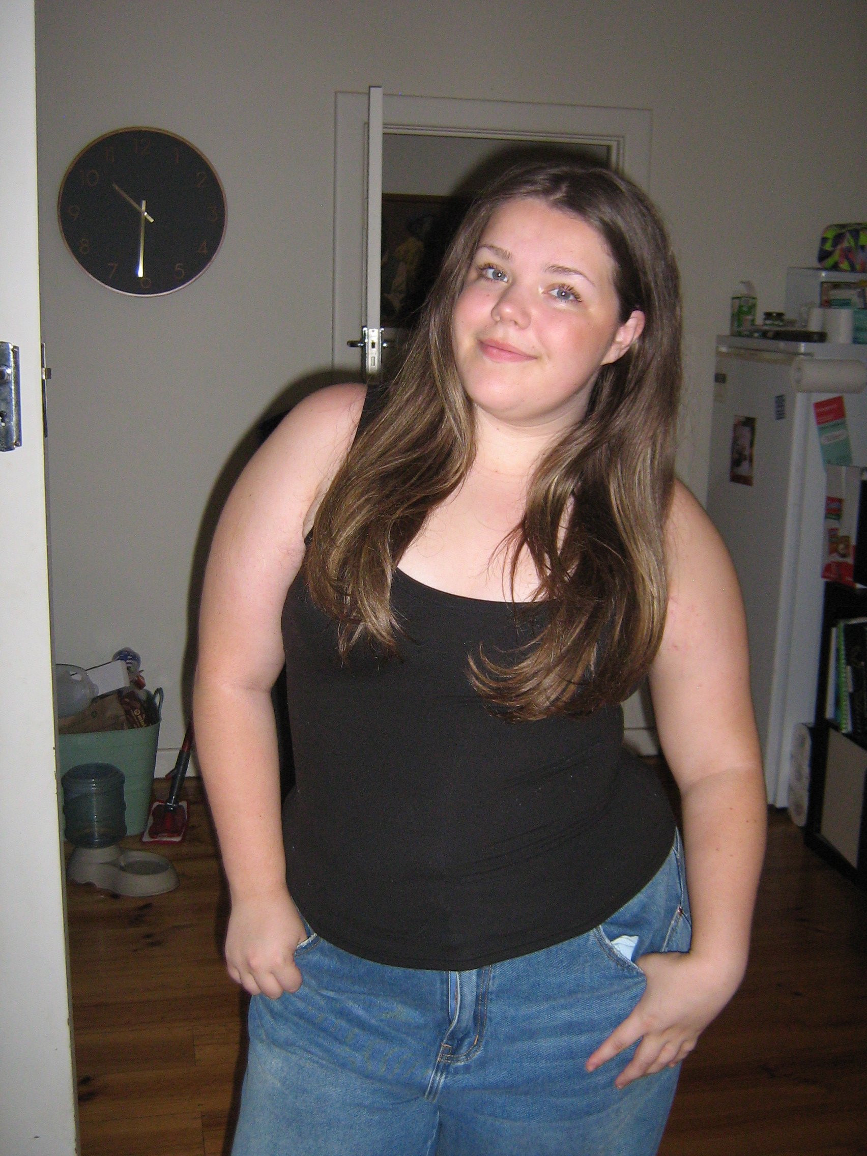 UGC and content creator  A young woman with long brown hair, wearing a black sleeveless top and blue jeans, standing in a kitchen, smiling and posing for a photo.