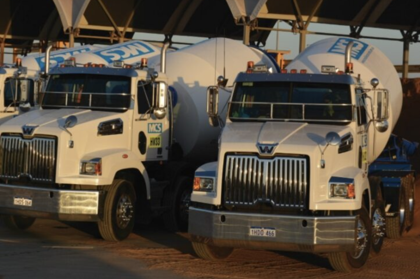 Two white cement tanker trucks parked side by side on a dirt surface, with large cylindrical tanks mounted on the back of each truck, under a metal roof structure.