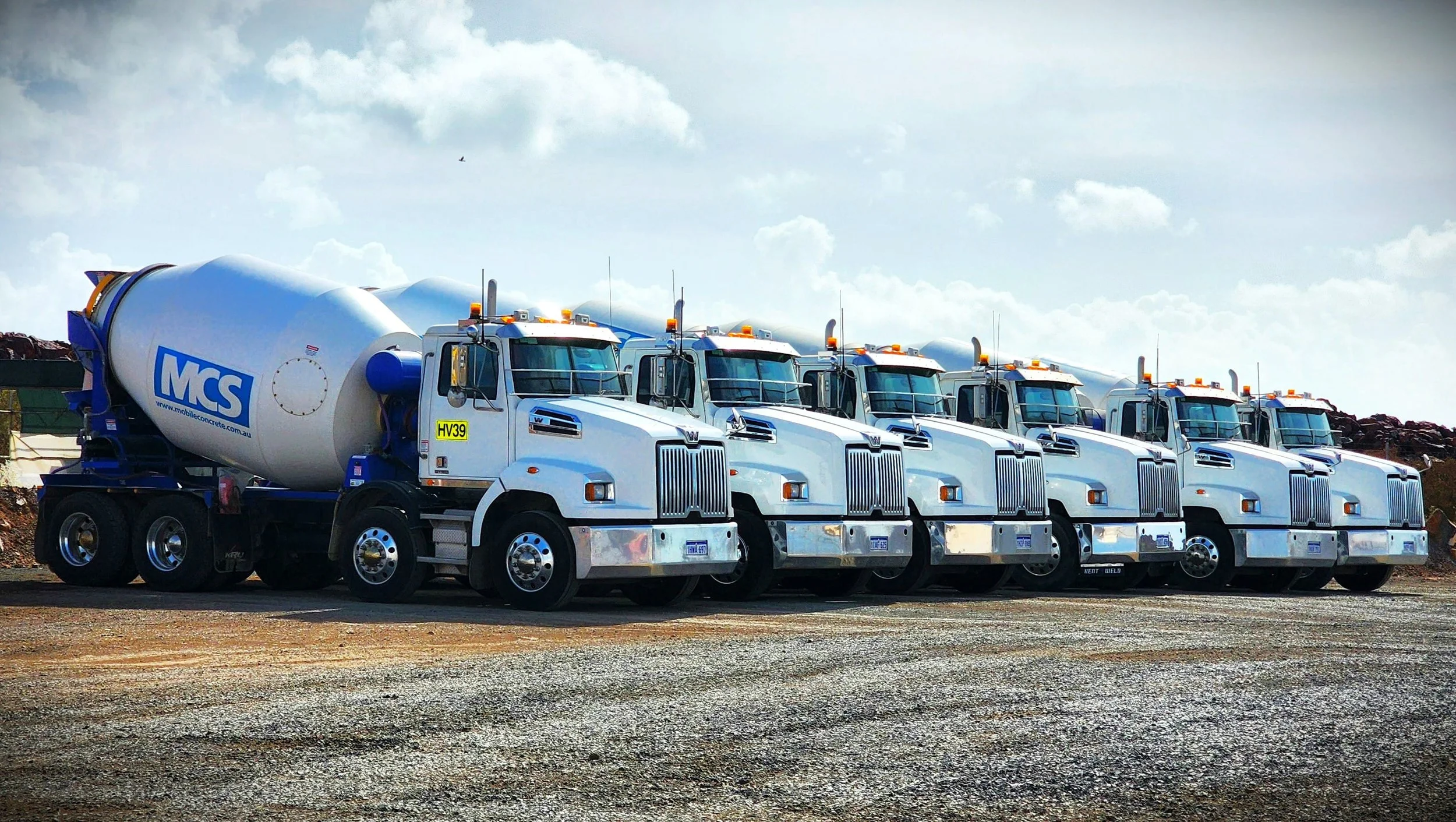 A line of six white cement mixer trucks parked outdoors on a gravel surface under a partly cloudy sky. The trucks have the logo MCS and a website URL on the mixing drums.