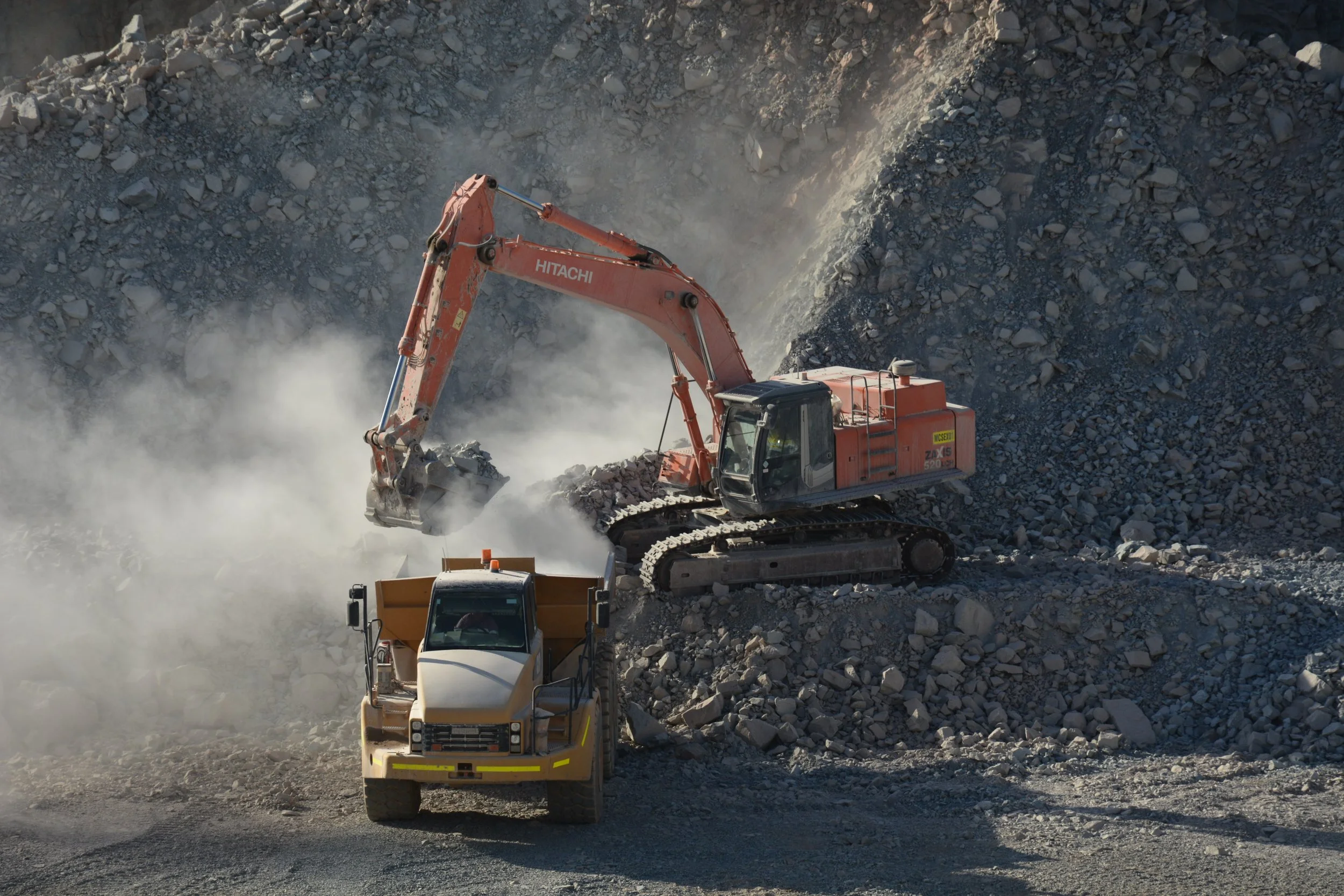 Construction site with excavator moving rocks and dust, truck and bulldozer working on dirt and rubble.
