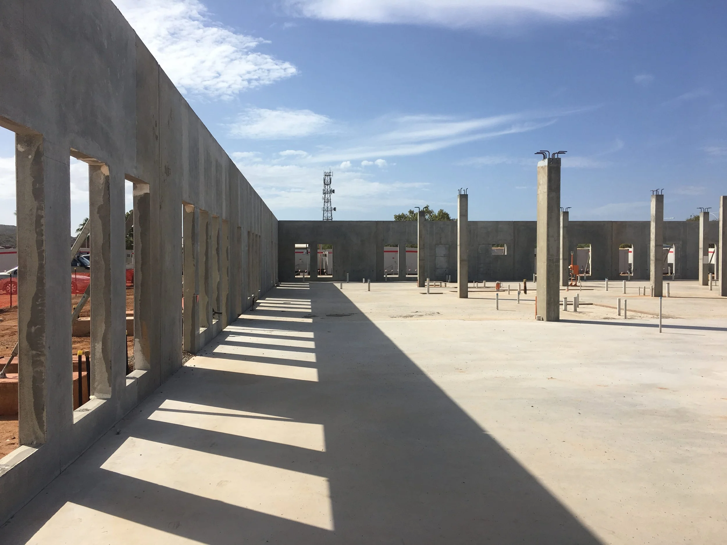 Construction site with concrete walls and tall pillars casting shadows, under a partly cloudy blue sky.