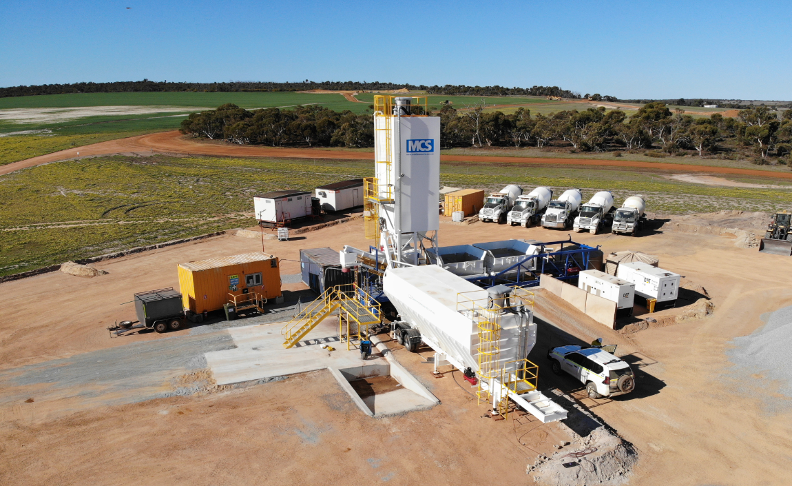 Construction site with equipment including cement mixers, trucks, and a large white tower with a blue logo, set in a rural landscape under a clear blue sky.