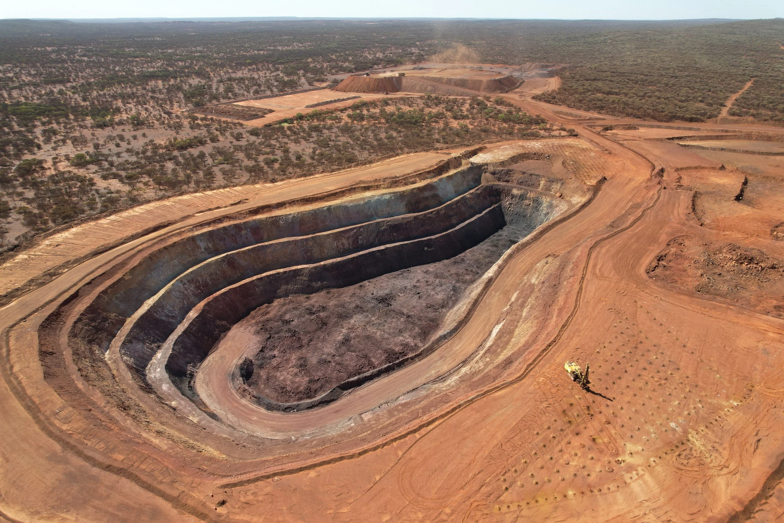 Aerial view of an open-pit mine in a flat, arid landscape with sparse vegetation. The mine has multiple terraced levels.