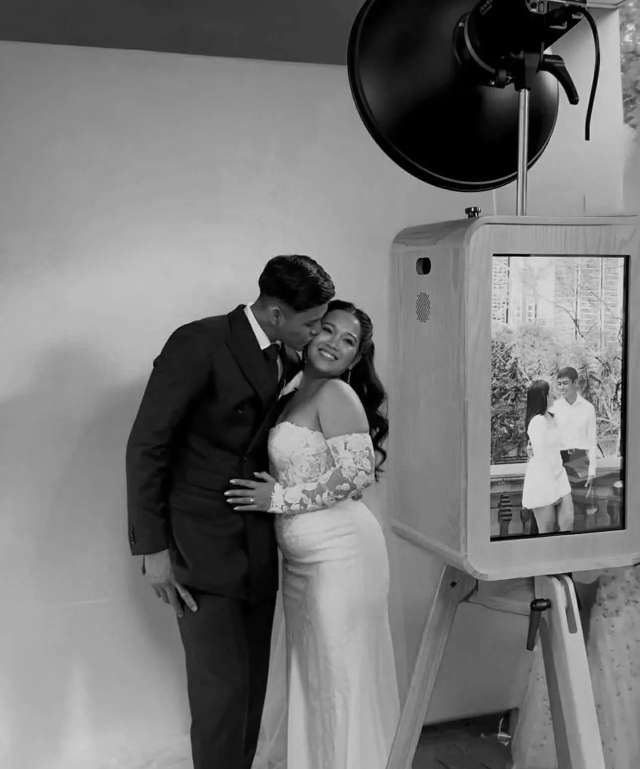A black and white photo of a bride and groom posing indoors, with a large photo booth screen showing their picture in the background. The groom is in a suit, kissing the bride on the cheek, who is smiling in a lace wedding dress.
