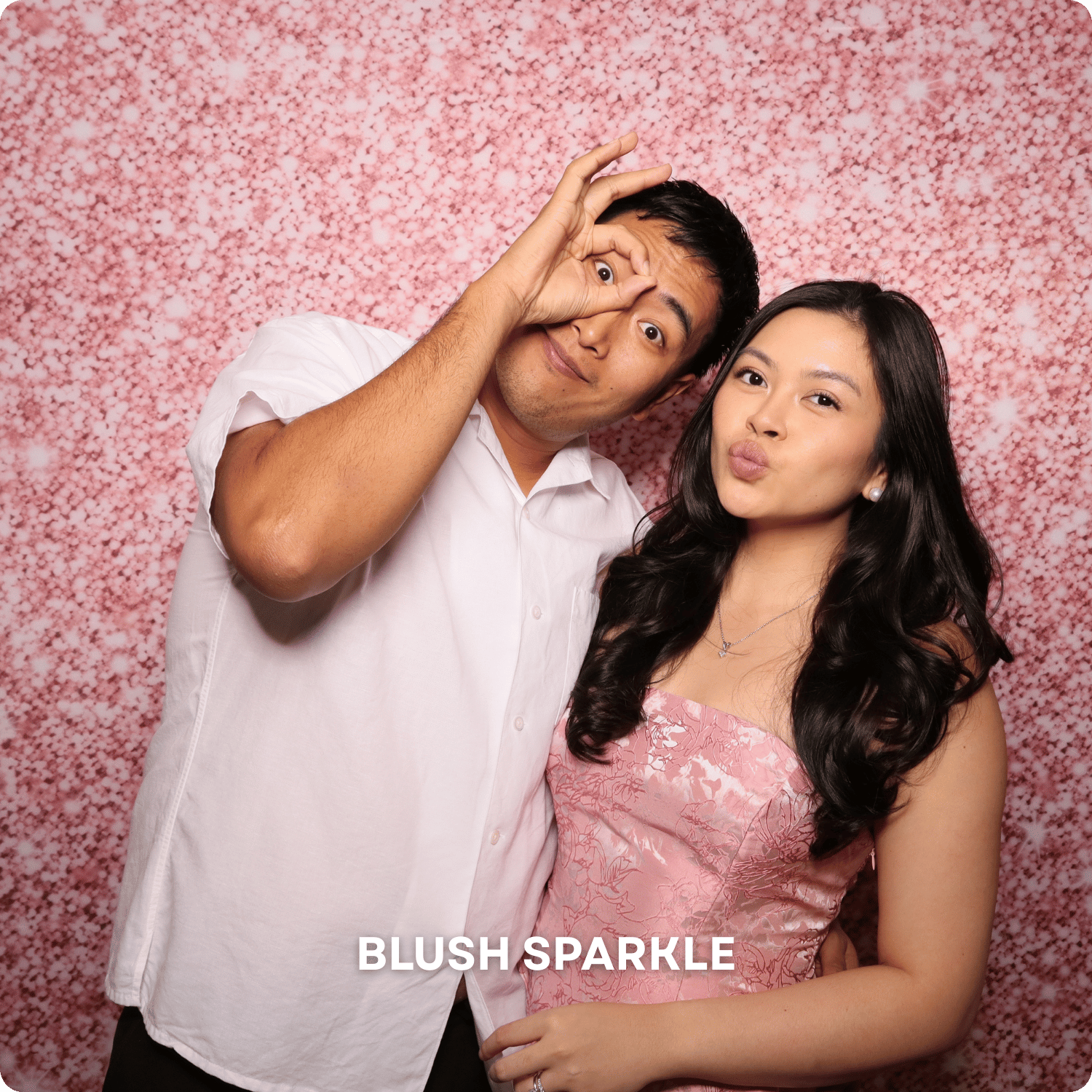A man and woman posing in front of a pink, sparkly backdrop. The man is making a circle with his fingers around his eye, and the woman is puckering her lips. The words 'BLUSH SPARKLE' are written at the bottom of the image.