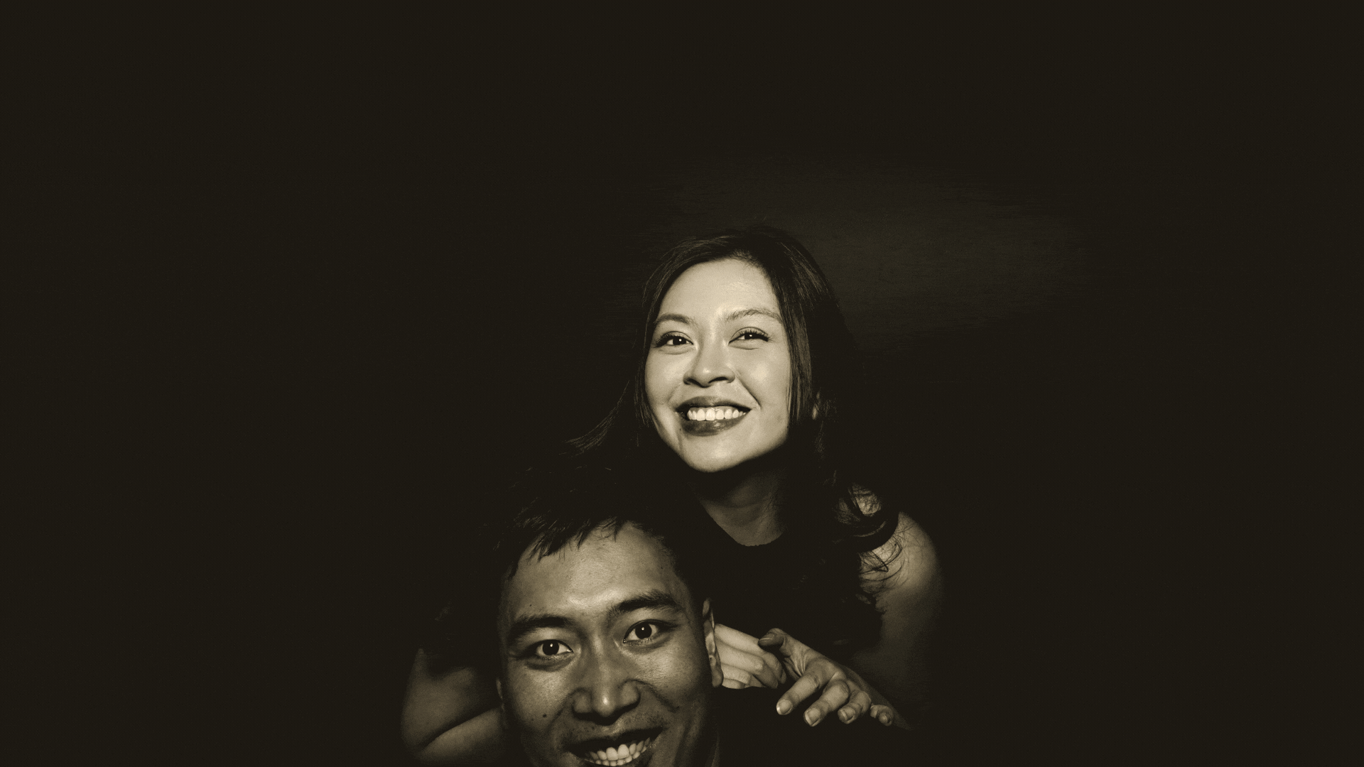 A black and white photo of an Asian woman smiling while sitting on the shoulders of a man, both looking at the camera, with a dark background.